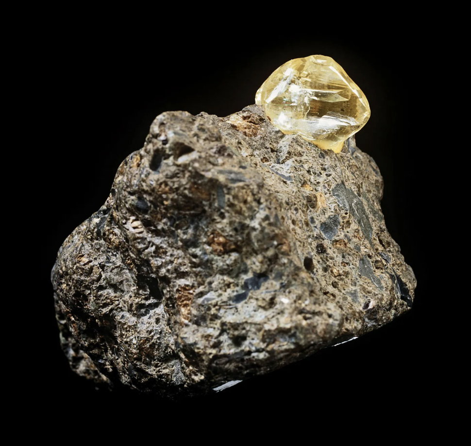 A rough, dark gray to black rock with a yellow, faceted gemstone on top against a black background.