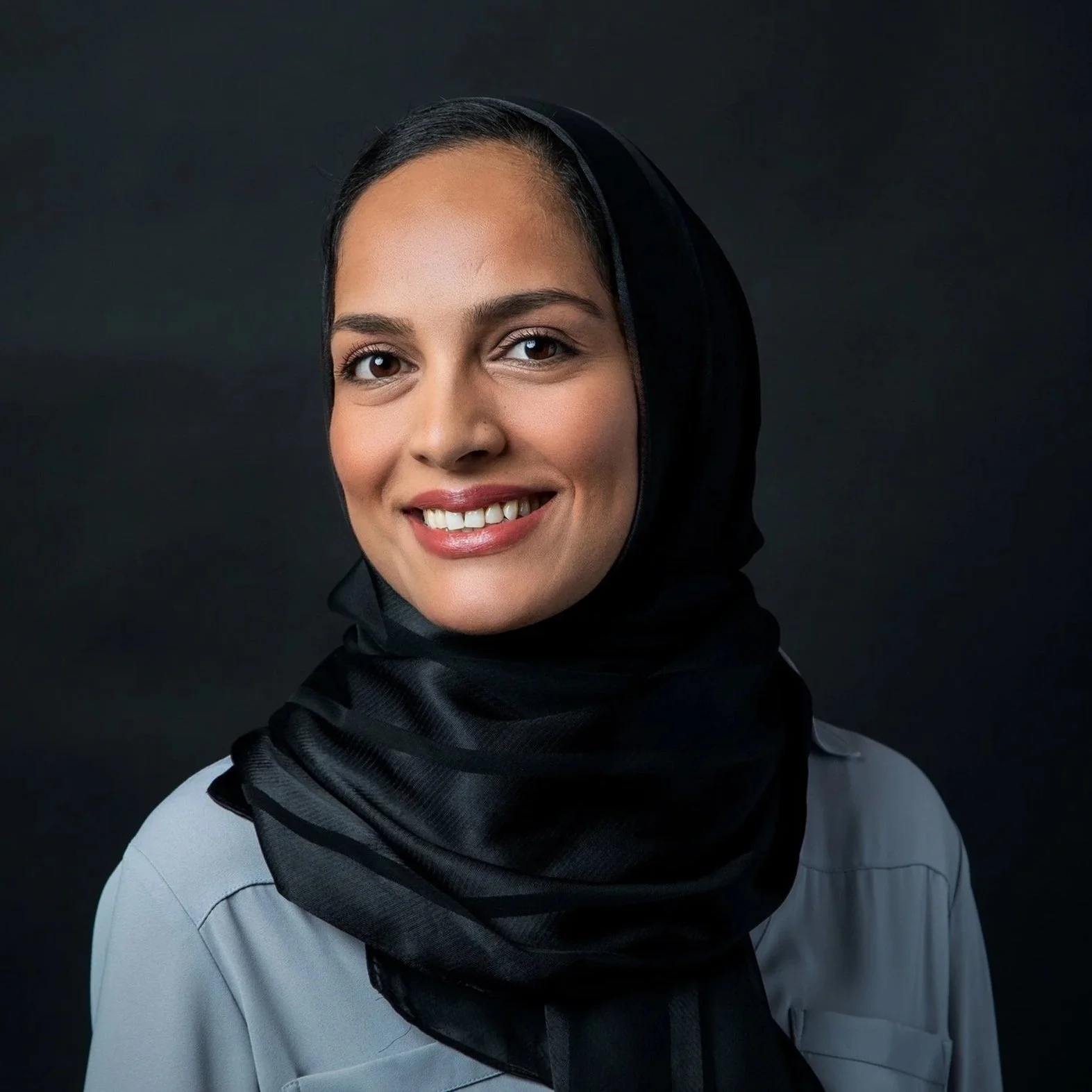 A woman with dark hair wearing a black hijab and a light gray blouse, smiling against a dark background.