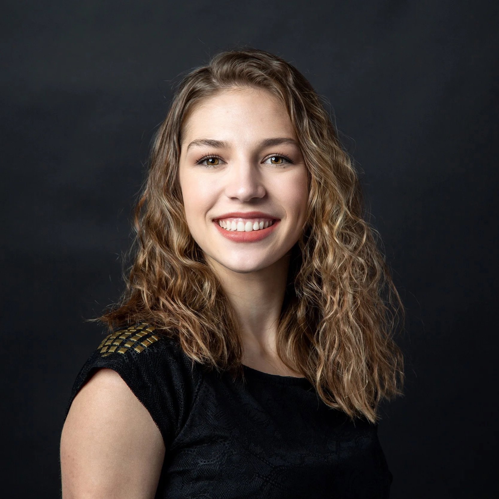 A young woman with curly light brown hair smiling at the camera, wearing a black top with gold accents on the shoulder, against a dark background.