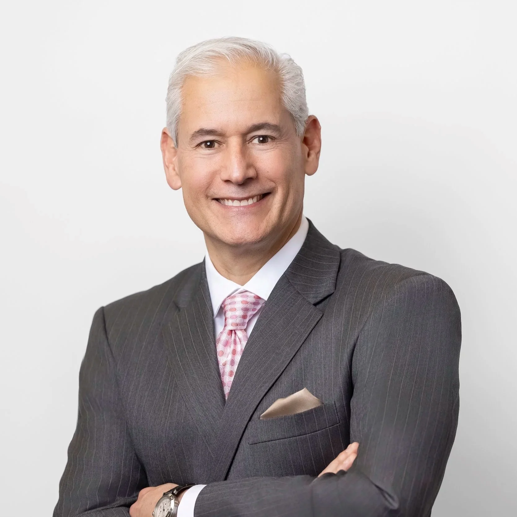 Portrait of a smiling middle-aged man with gray hair, wearing a dark gray pinstripe suit, white shirt, pink patterned tie, and a beige pocket square, standing with arms crossed against a plain white background.