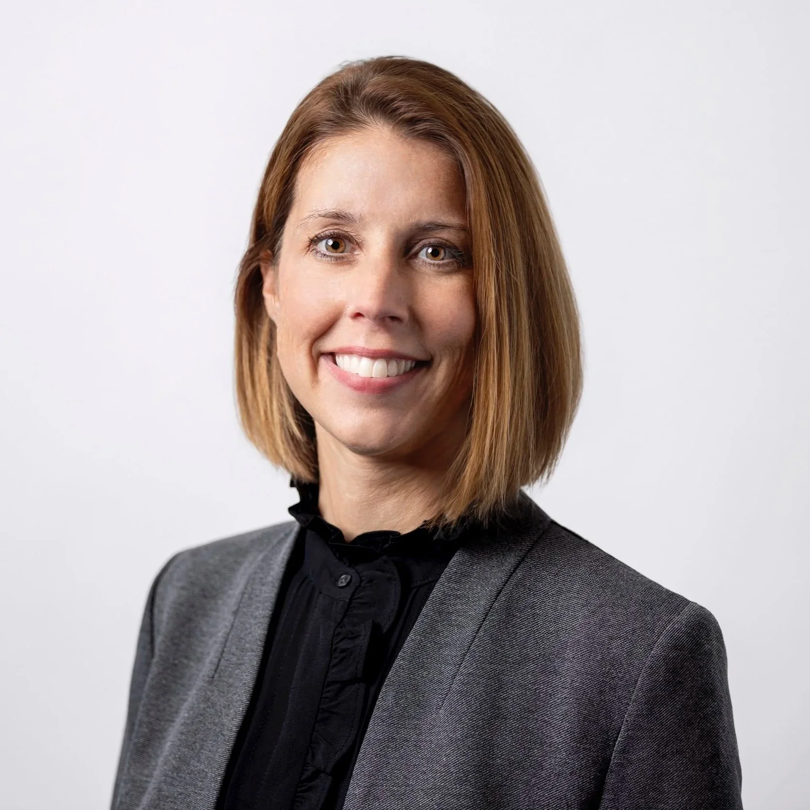Portrait of a professional woman with shoulder-length light brown hair, smiling, wearing a dark blouse and gray blazer, against a plain white background.