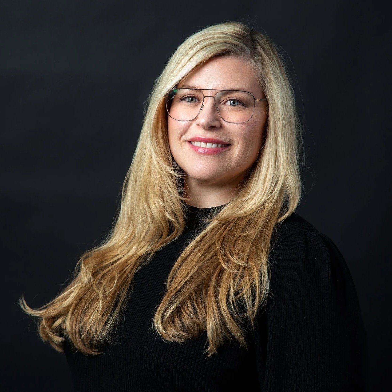 A woman with long blonde hair wearing glasses and a black top, smiling against a dark background.