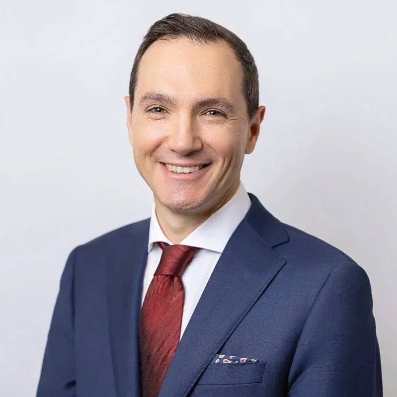 Professional portrait of a smiling man in a navy suit, white shirt, and red tie against a plain background.