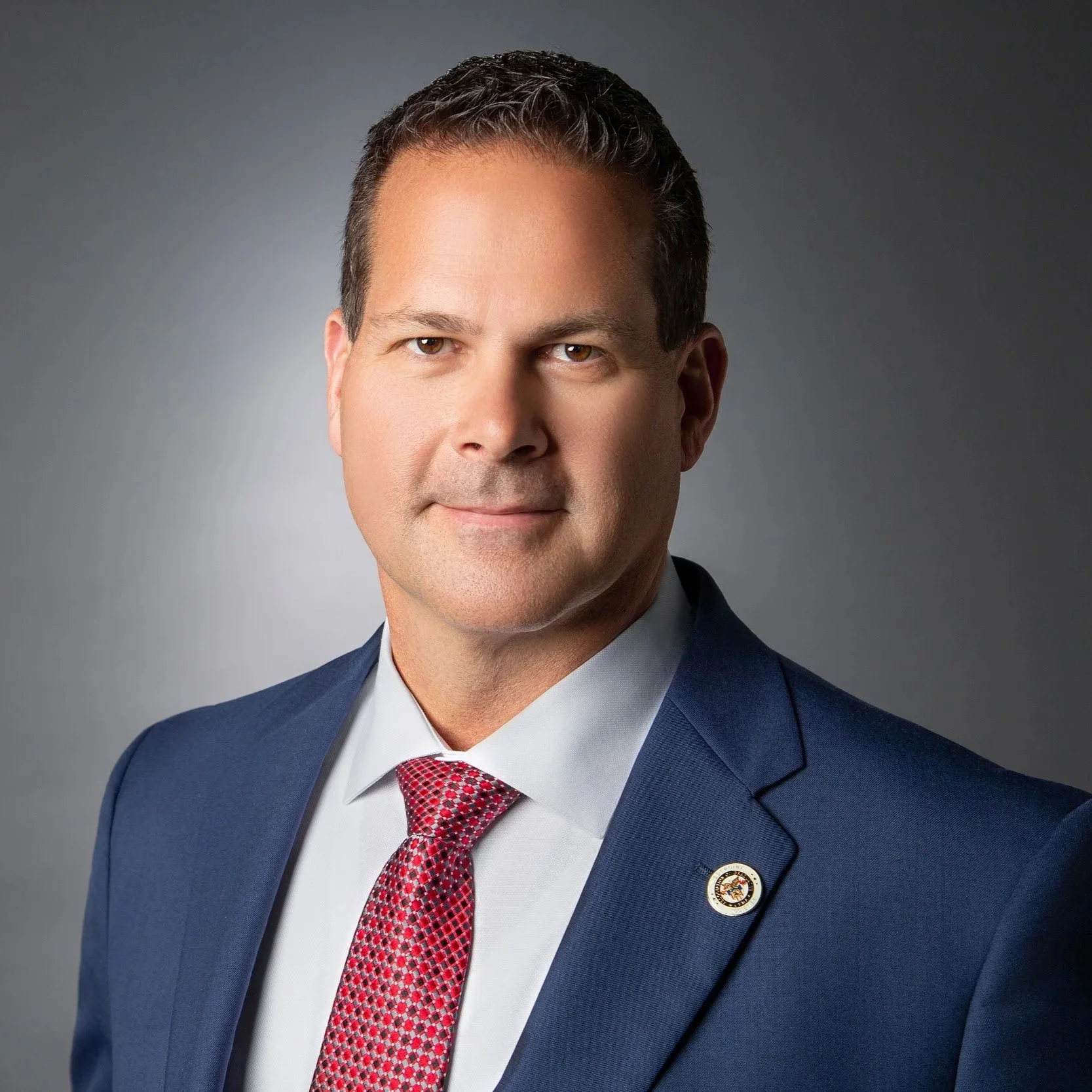 Headshot of a man in a navy suit, white shirt, and red patterned tie, against a gray background.