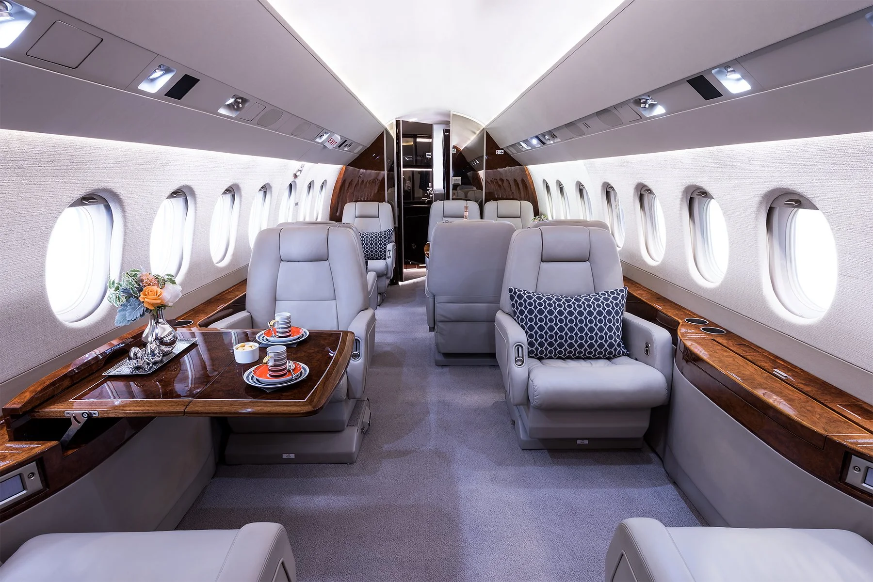 Interior of a private jet with plush gray seats, wooden side tables, windows with white curtains, and a small table set with cups and flowers.