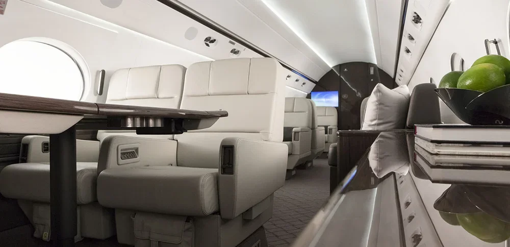 Interior of a private jet with white leather seats, a small table, and a bar area with green apples and magazines.
