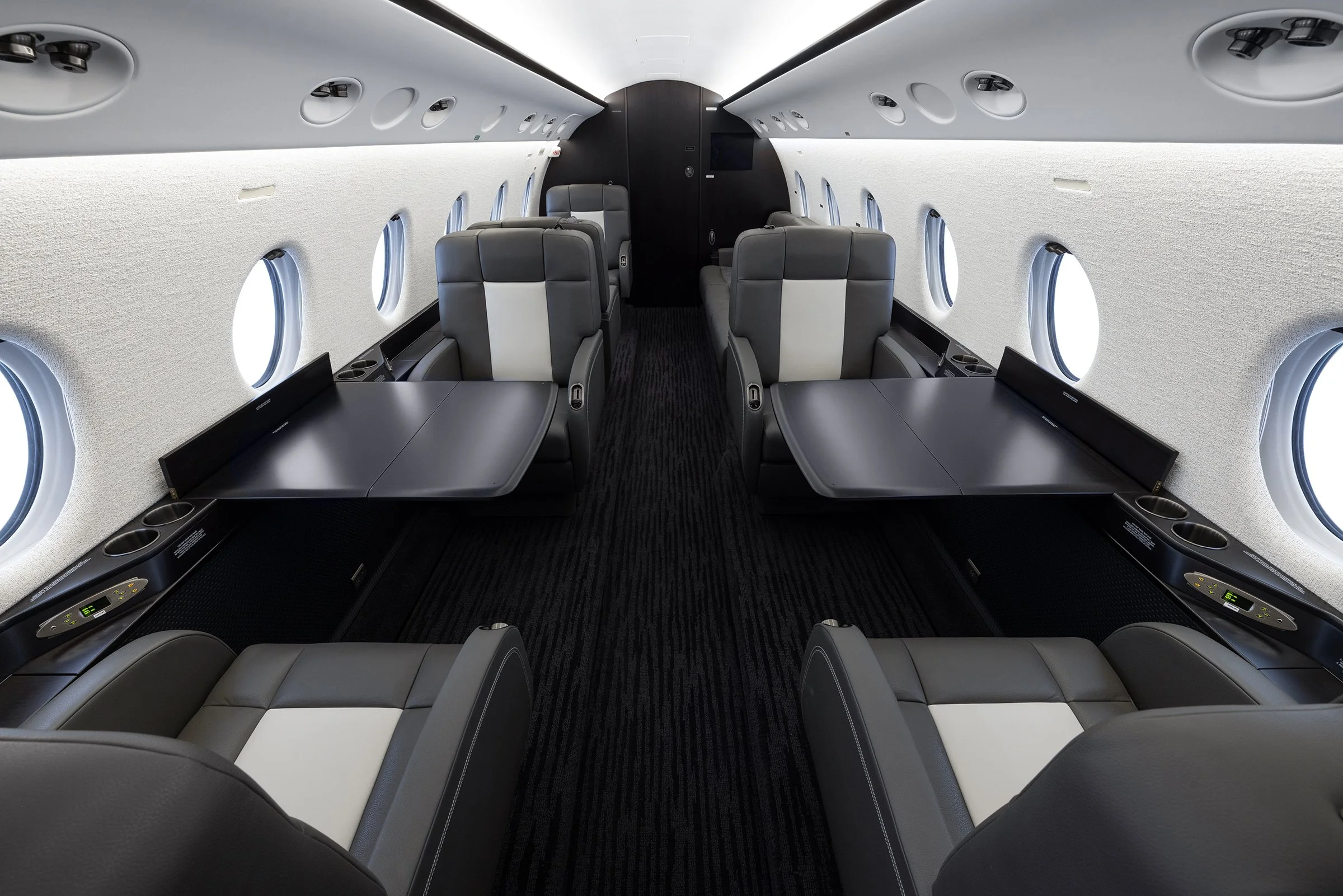 The interior of a modern private jet with luxurious gray leather seats, large windows, and built-in tables.
