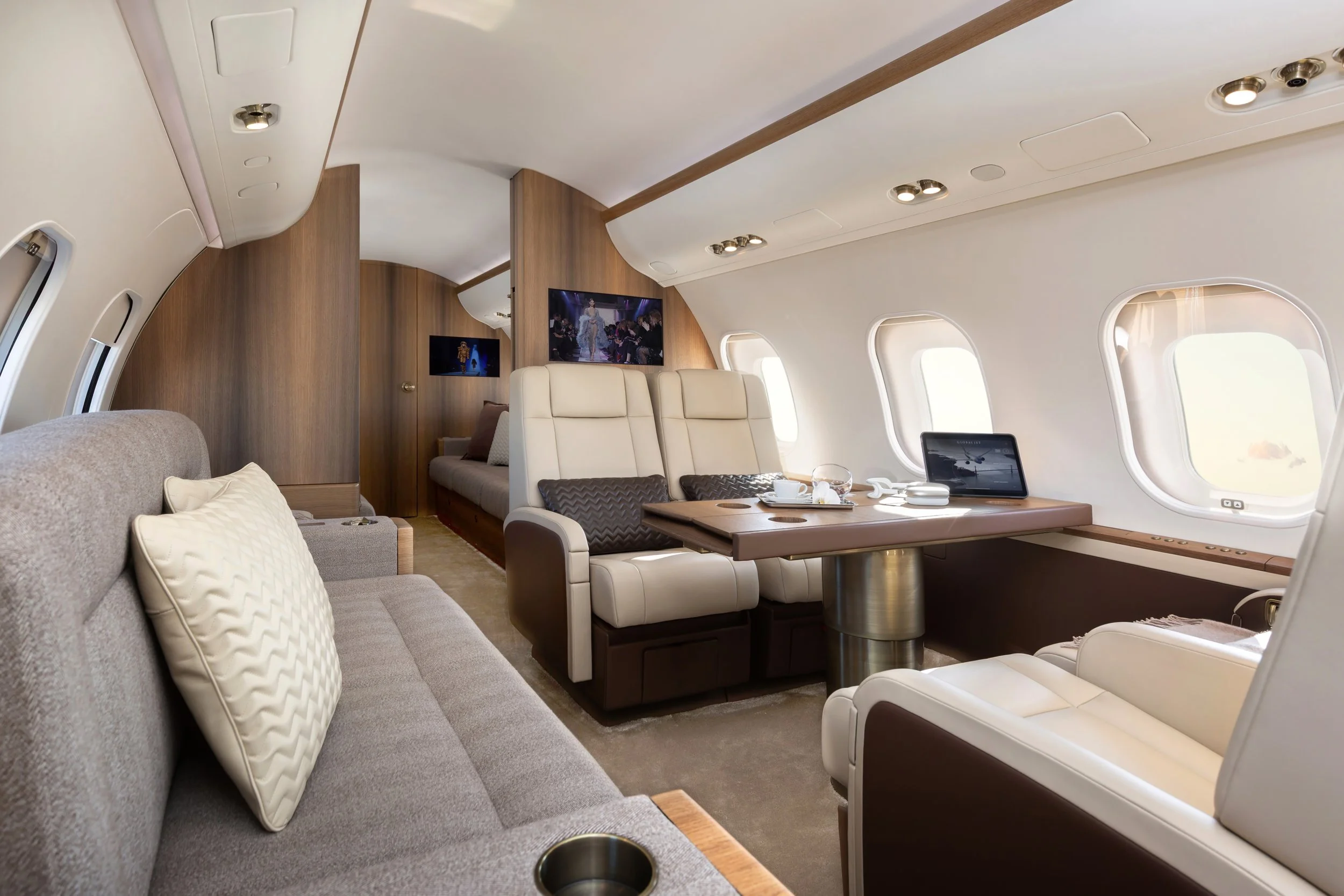 Interior of a luxury airplane cabin with beige and brown leather seats, a wooden table set with cups and a tablet, and windows showing the sky outside.