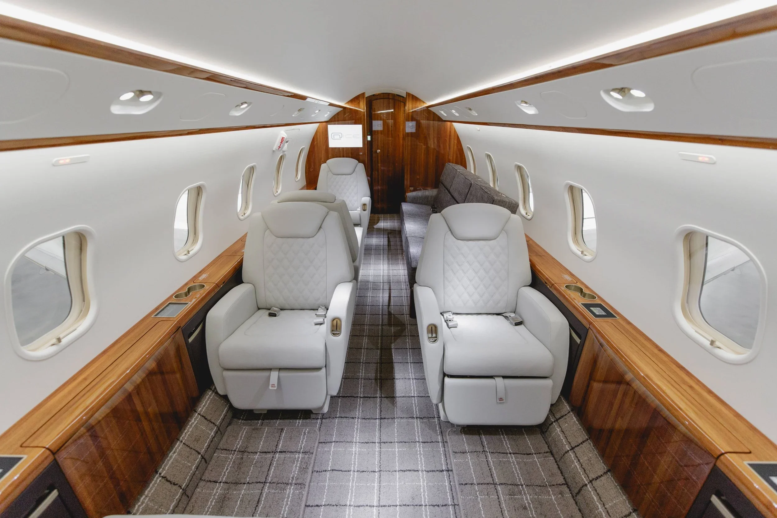 The interior of a private jet with white leather seats, a gray sofa, and wooden accents.
