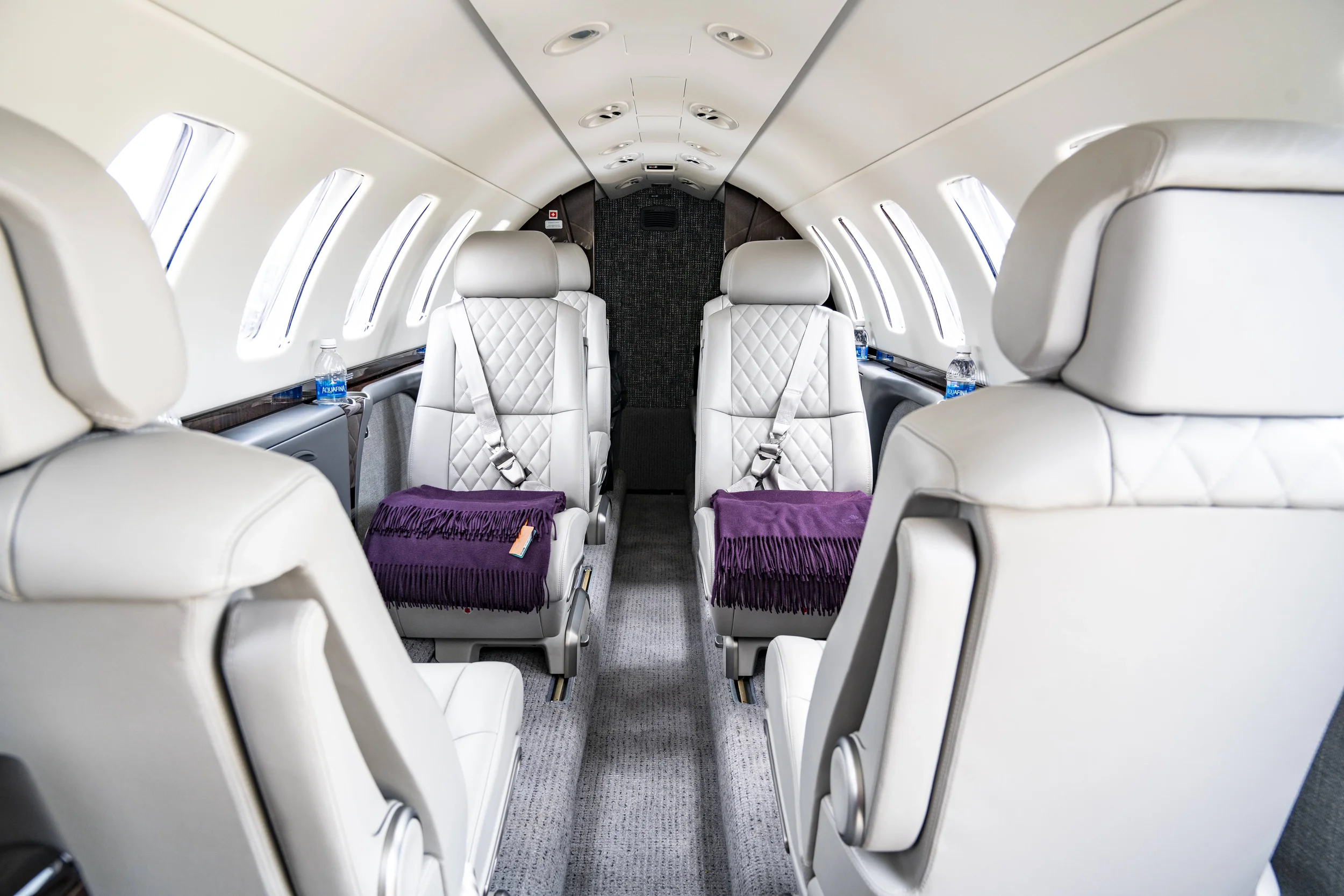 Interior of a private jet with white leather seats, purple blankets, and water bottles on the side compartments.