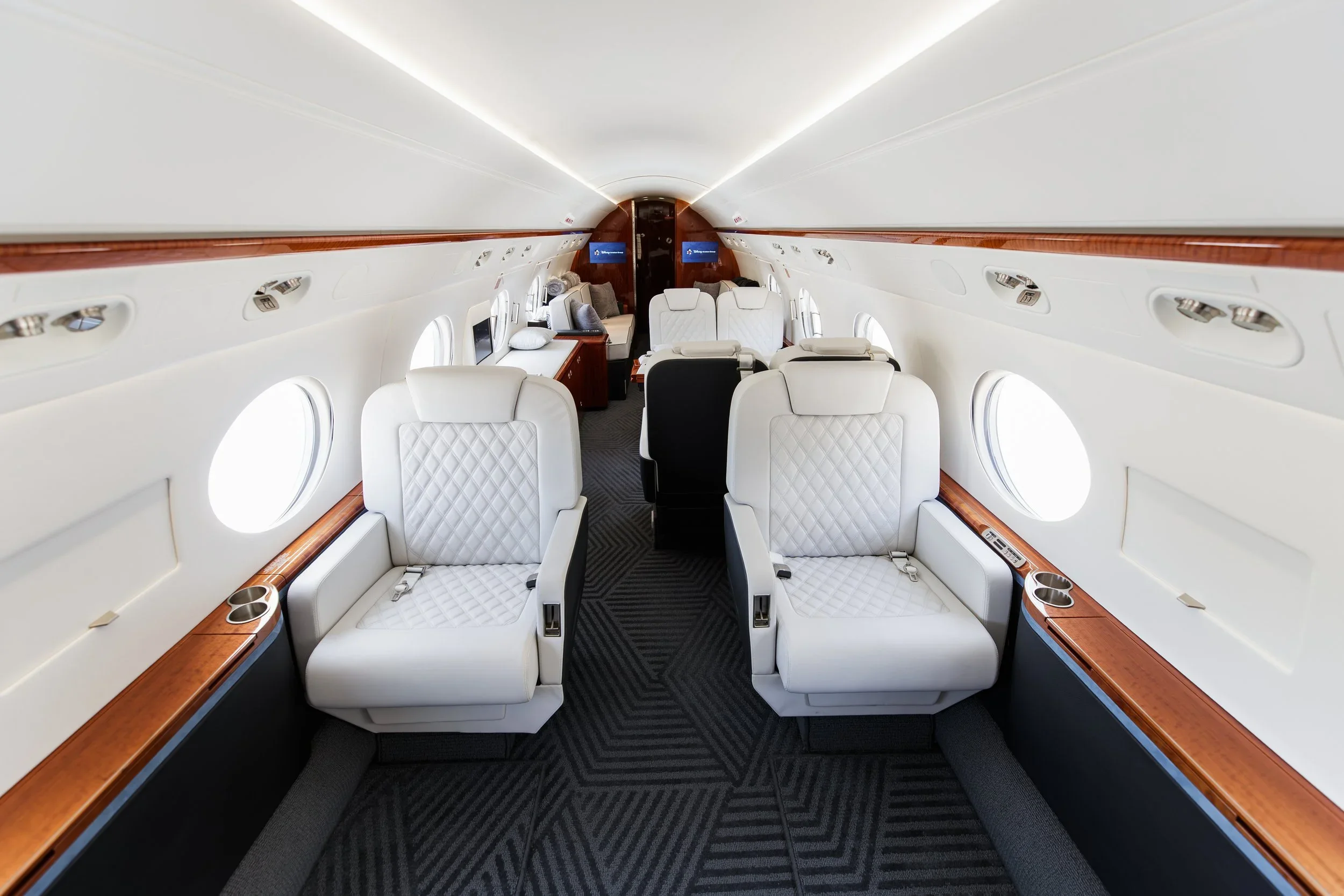 Interior of a private jet with white leather seats, wood accents, and windows along the sides.