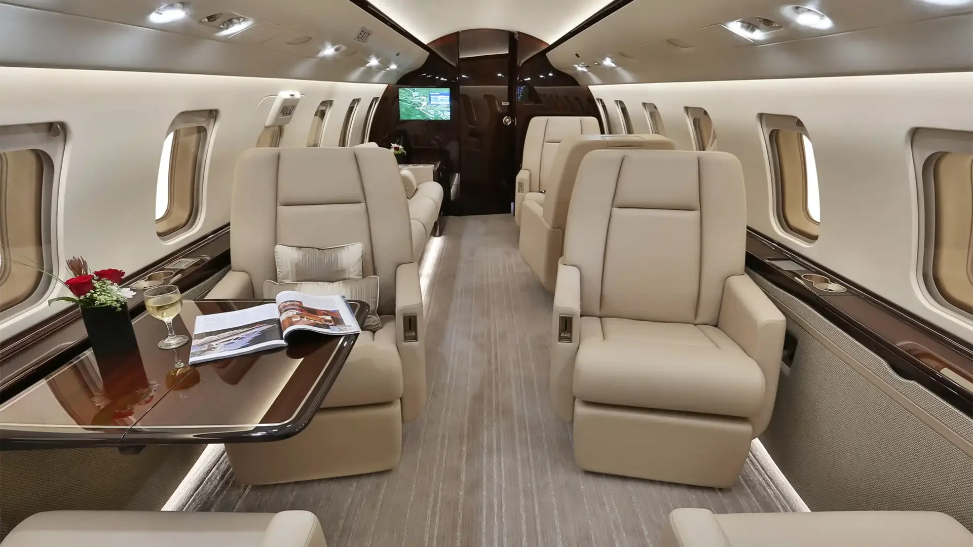 Inside a luxurious private jet with beige leather seats, a wooden table with a bouquet of red flowers, a glass of white wine, and an open magazine, overlooking windows with blinds.