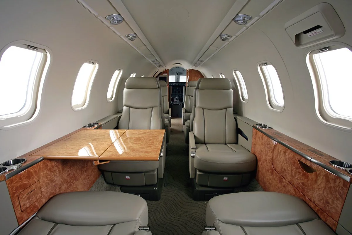 Interior of a private jet with beige leather seats, small side tables with cup holders, and windows along the cabin walls.