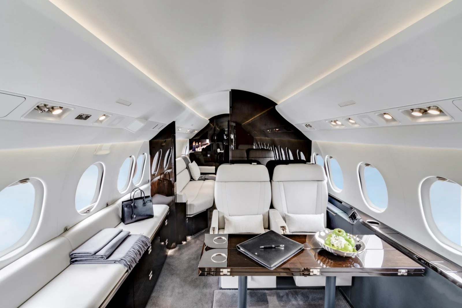 Interior of a private jet with cream leather seats, a wooden table with a black folder and pen, and a bowl of green grapes, with windows showing a blue sky outside.