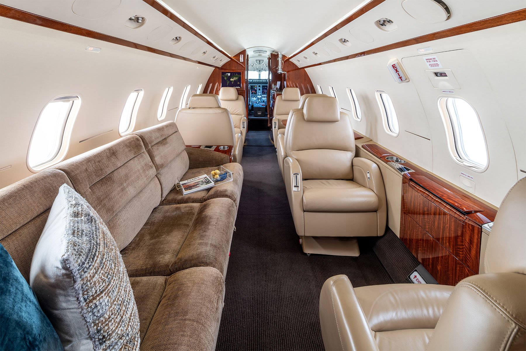 Inside the cabin of an airplane, showing beige leather seats, brown fabric sofa, windows along both sides, and wood paneling on the walls.