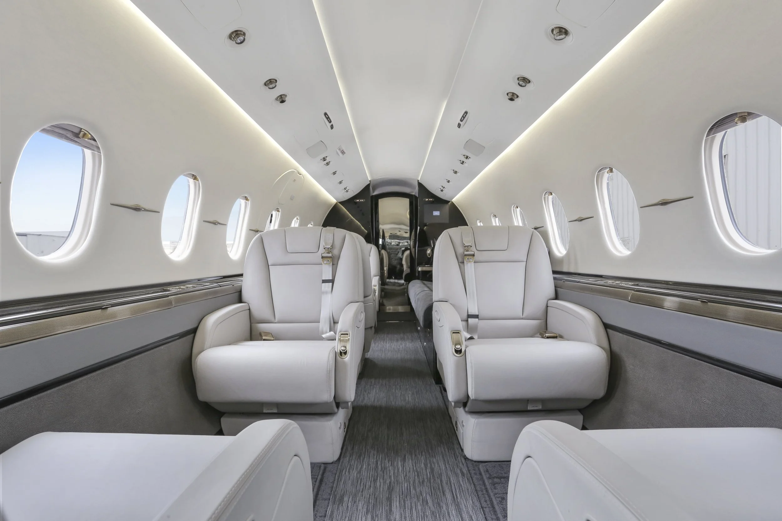 Interior of a private jet with white leather seats and several windows, looking towards the cockpit door.