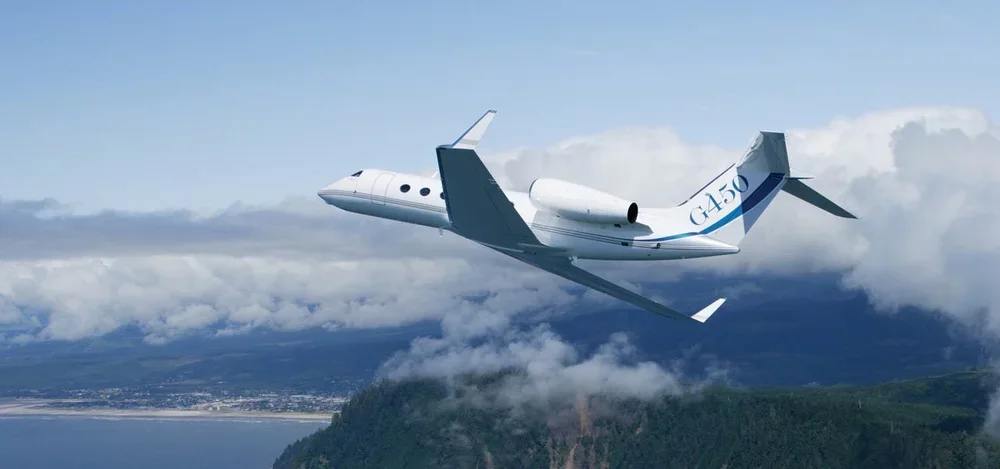 A white private jet with G450 written on the tail, flying high above green mountains and partly cloudy sky.