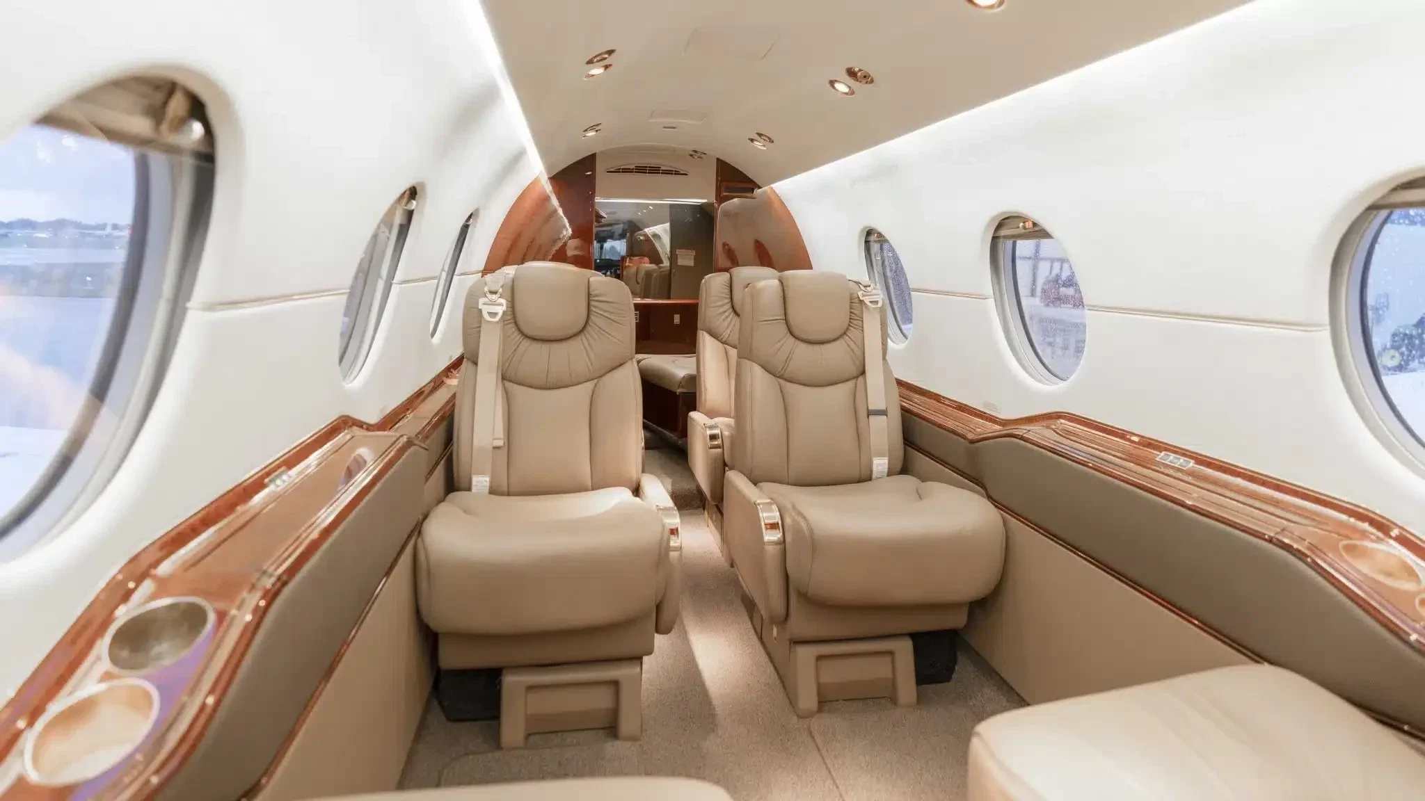 Interior of a private jet with beige leather seats, oval windows, and wooden trim.