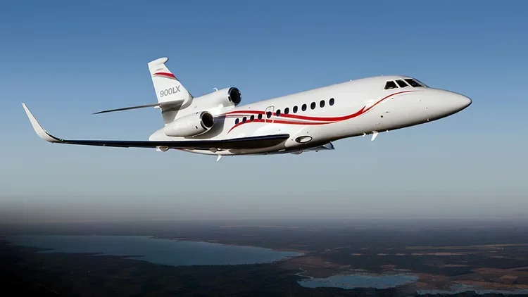 A white private jet with red and black stripes flying in a clear blue sky over a landscape with lakes and fields.