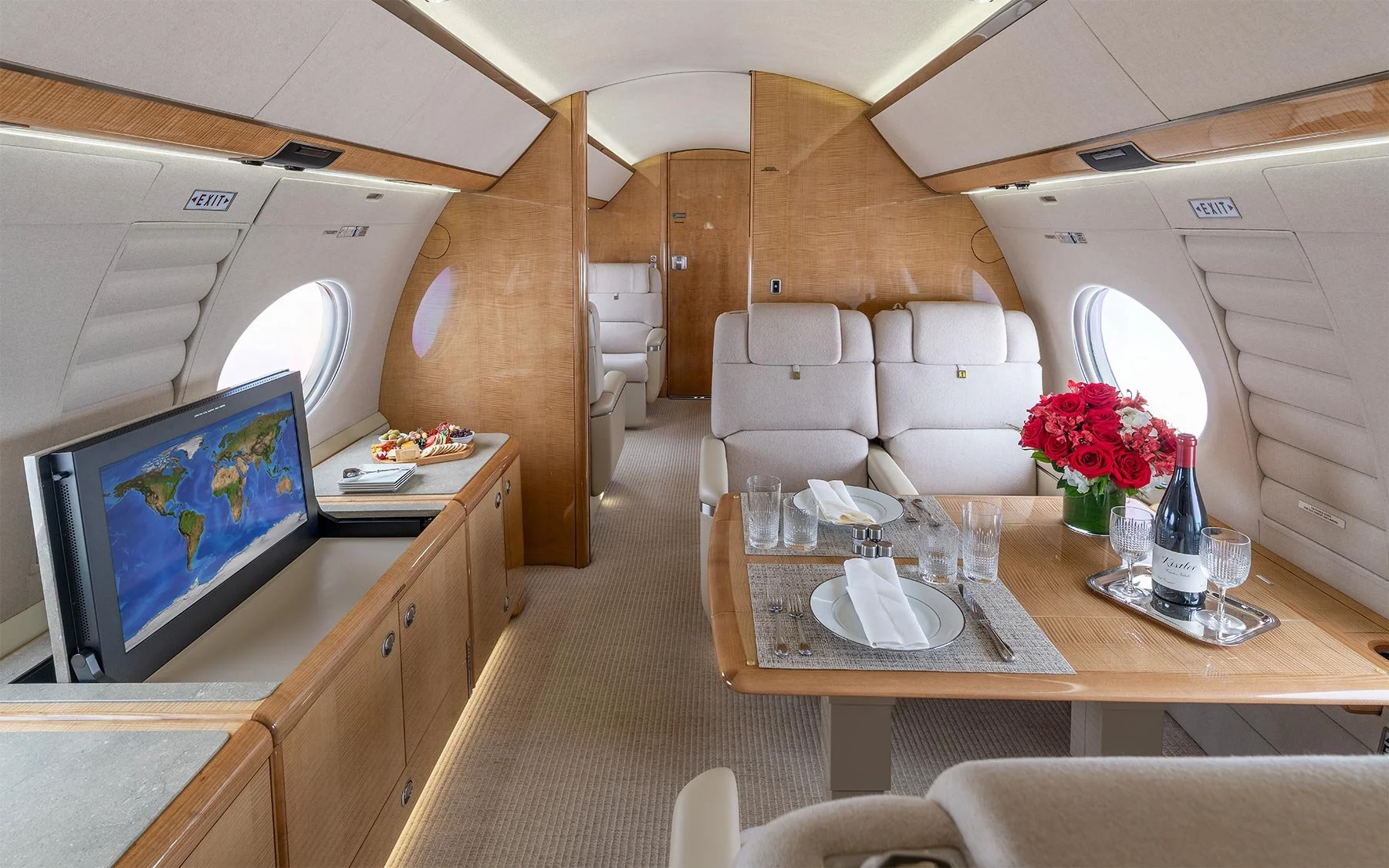 Luxurious airplane interior with plush seats, a wooden dining table set for a meal with wine and flowers, and a television showing a world map.