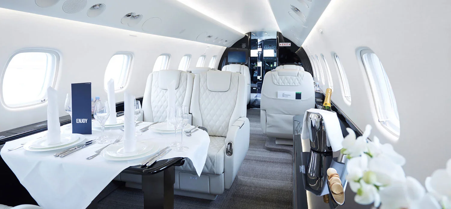 Inside a private jet with white leather seats, set dining table with glassware and utensils, and a champagne bottle on a side table.