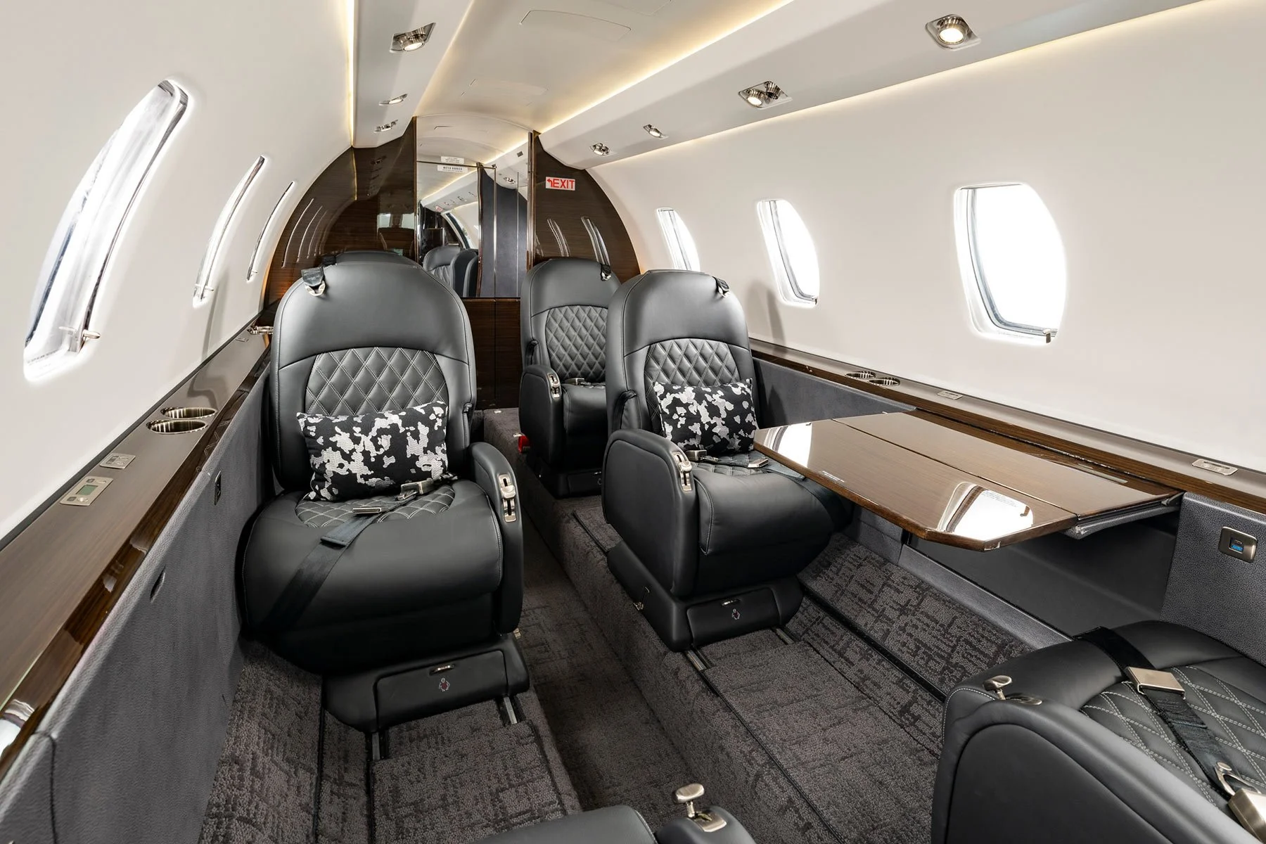 Interior of a private jet with four black leather seats, each with a patterned pillow, and a folding table along the window side.