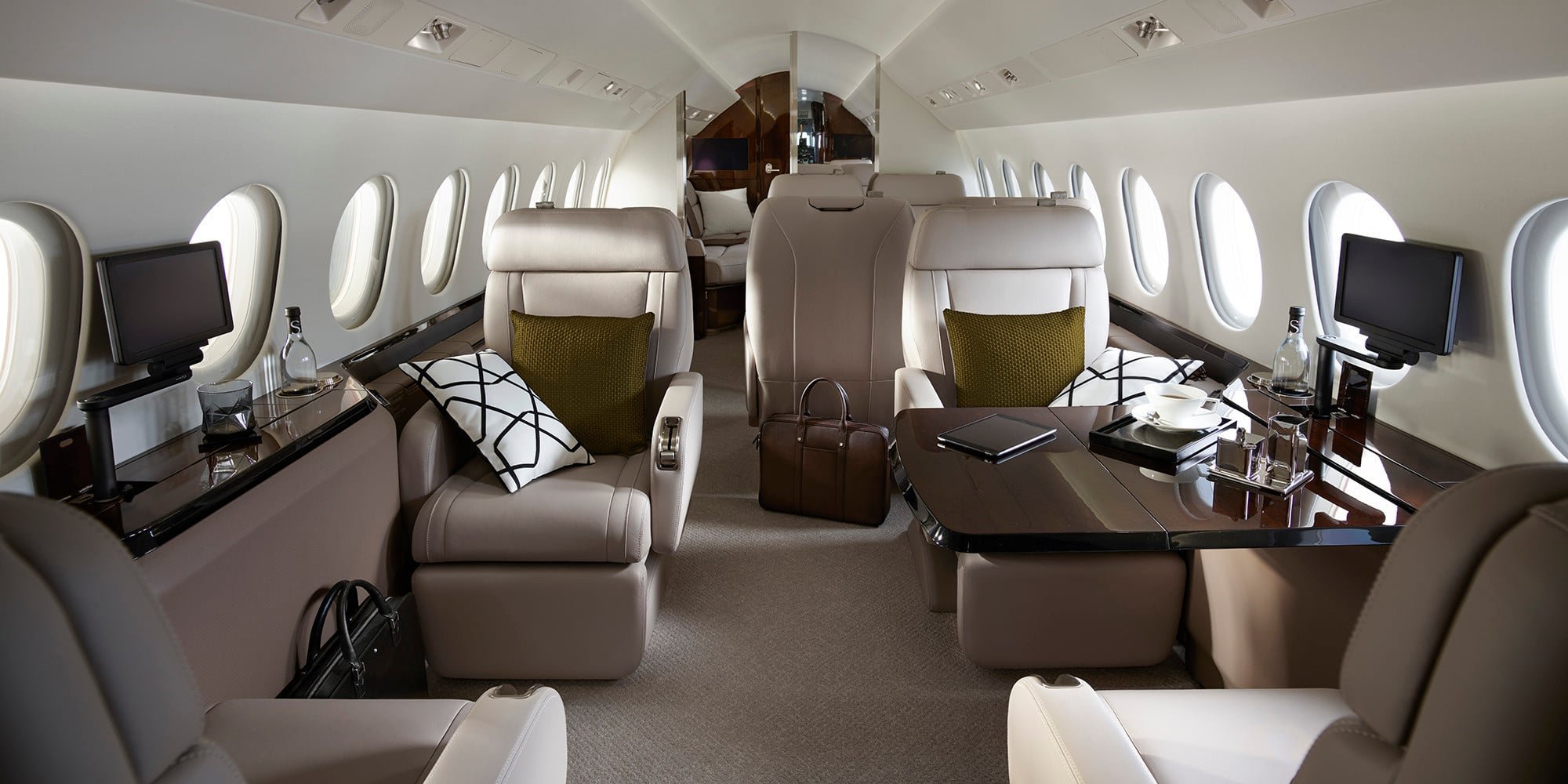 Luxury airplane interior with beige leather seats, small tables, two monitors, pillows, water bottles, and a brown leather briefcase.