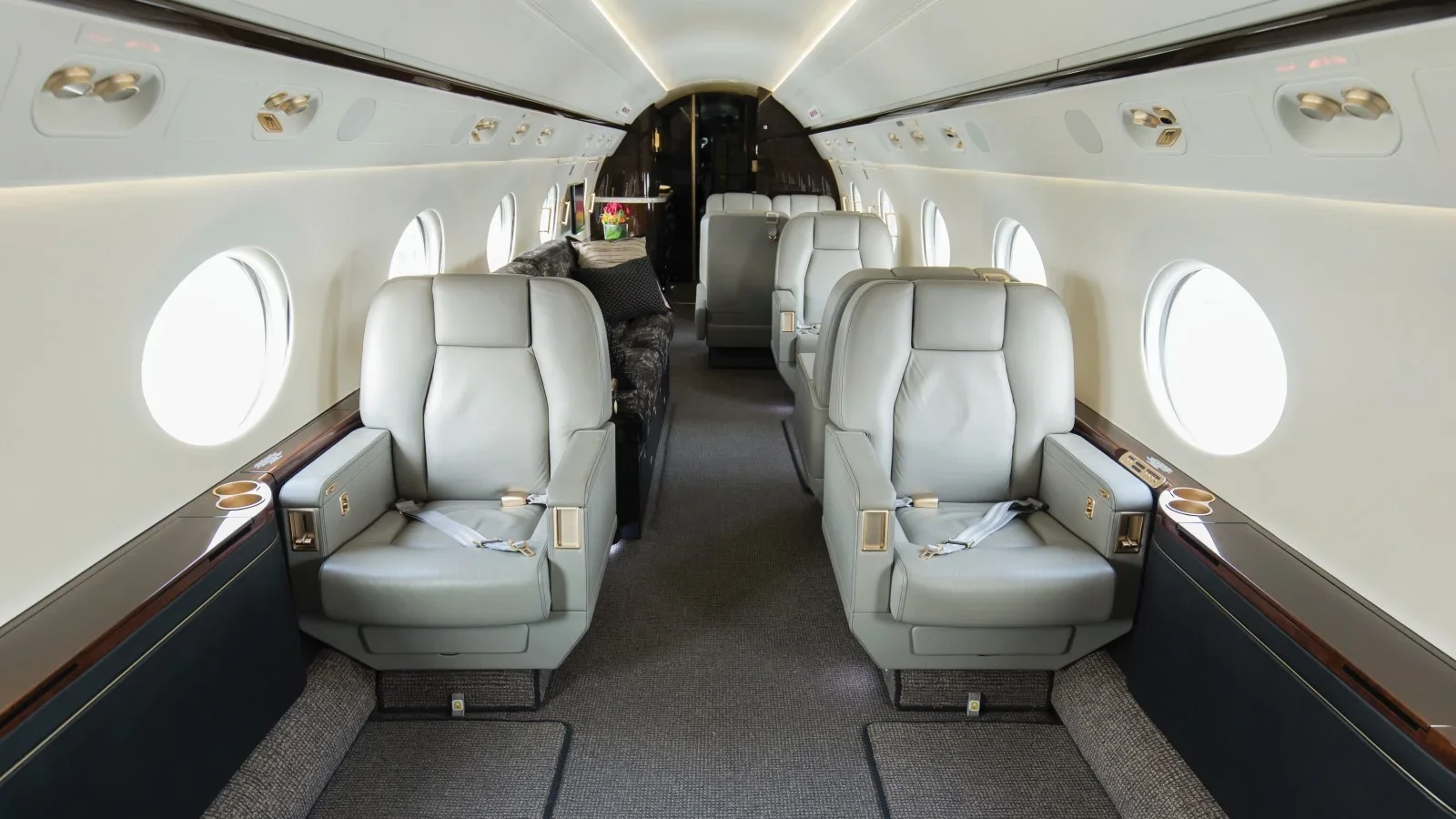 Interior of a private jet with white leather seats, windows on both sides, and a dark wood trim along the side panels. There are four seats visible, arranged in pairs facing each other, with a small table or storage area between them. A flower arrangement is at the front near the cockpit.