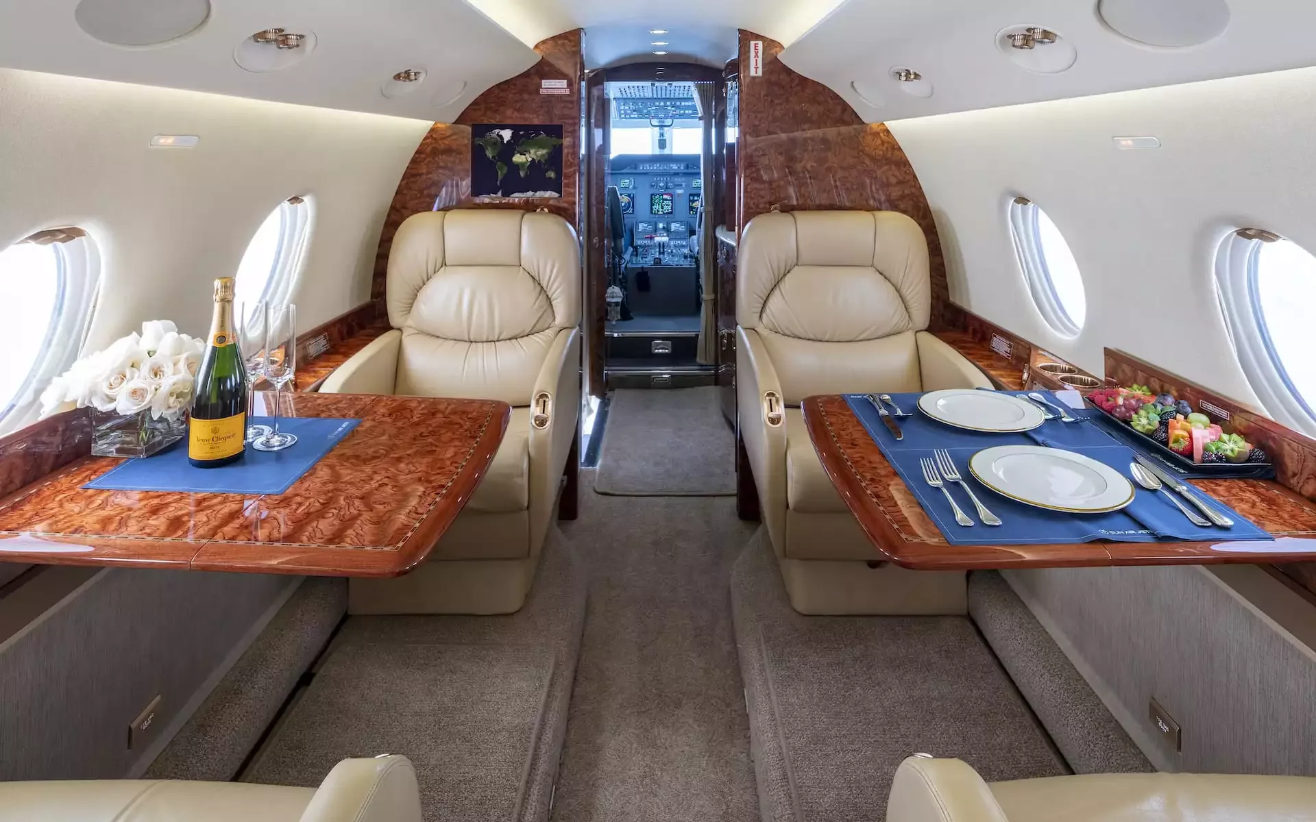 Interior of a private jet with two plush beige leather seats facing each other, a small table set with plates and cutlery, and a champagne bottle with glasses, with windows on each side and the cockpit visible in the background.