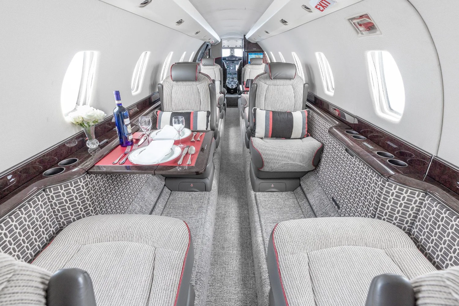Interior of a private jet with plush seats, a table set with wine glasses, a bottle of wine, and a floral centerpiece.