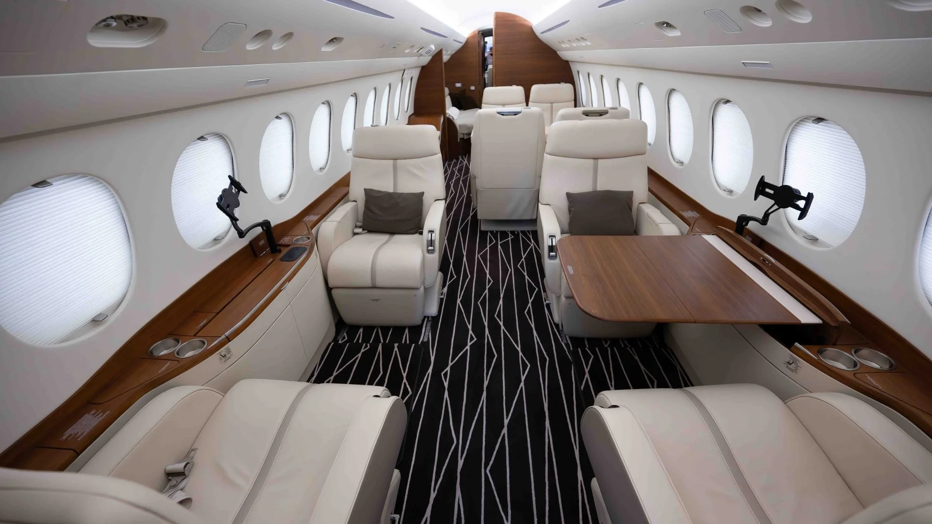 A luxurious private jet interior with cream-colored leather seats, large windows with shades, wooden accents, and a patterned black and white carpet.