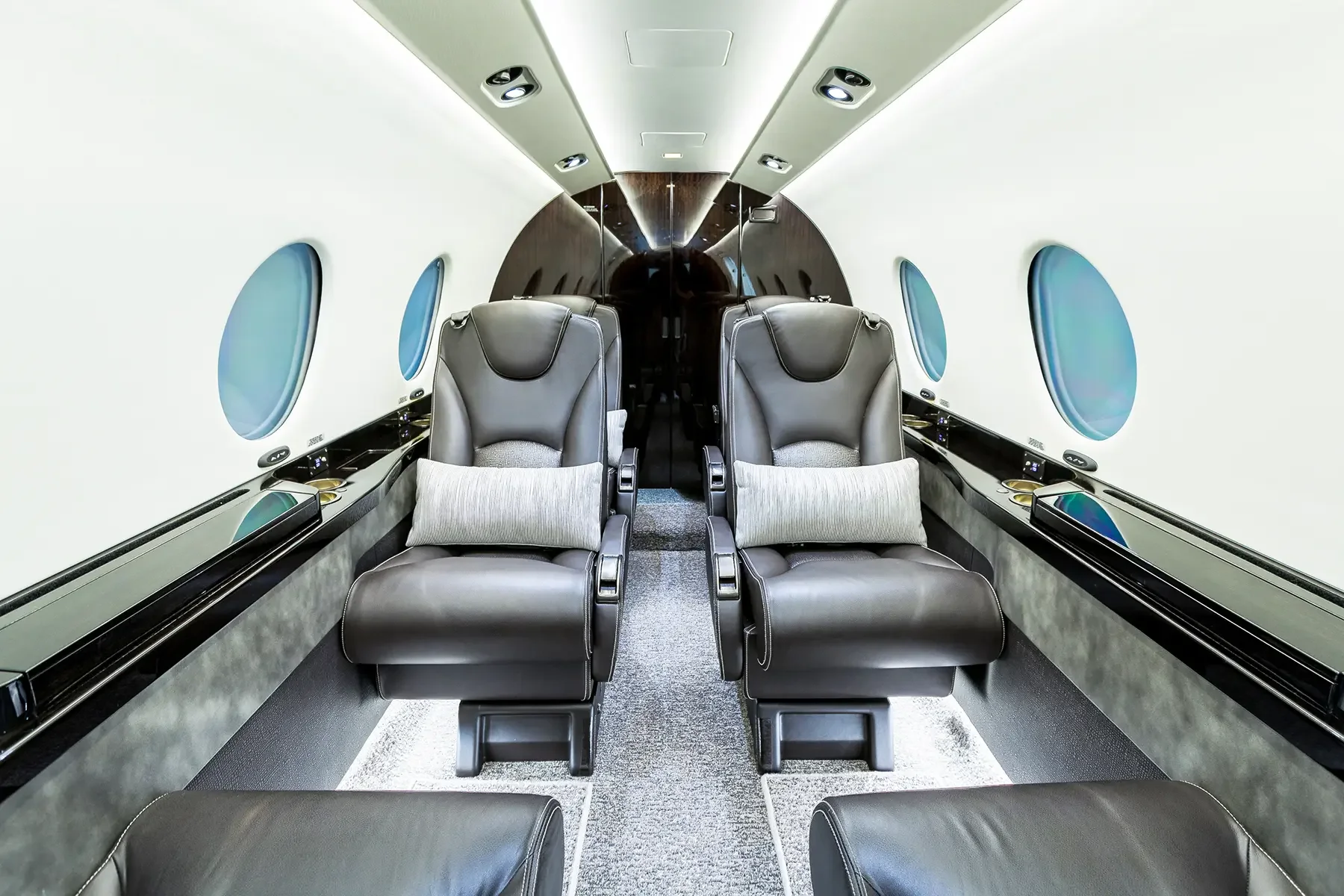 Inside a modern airplane, two leather seats with pillows, windows on the sides, and reflective panels on the wall.