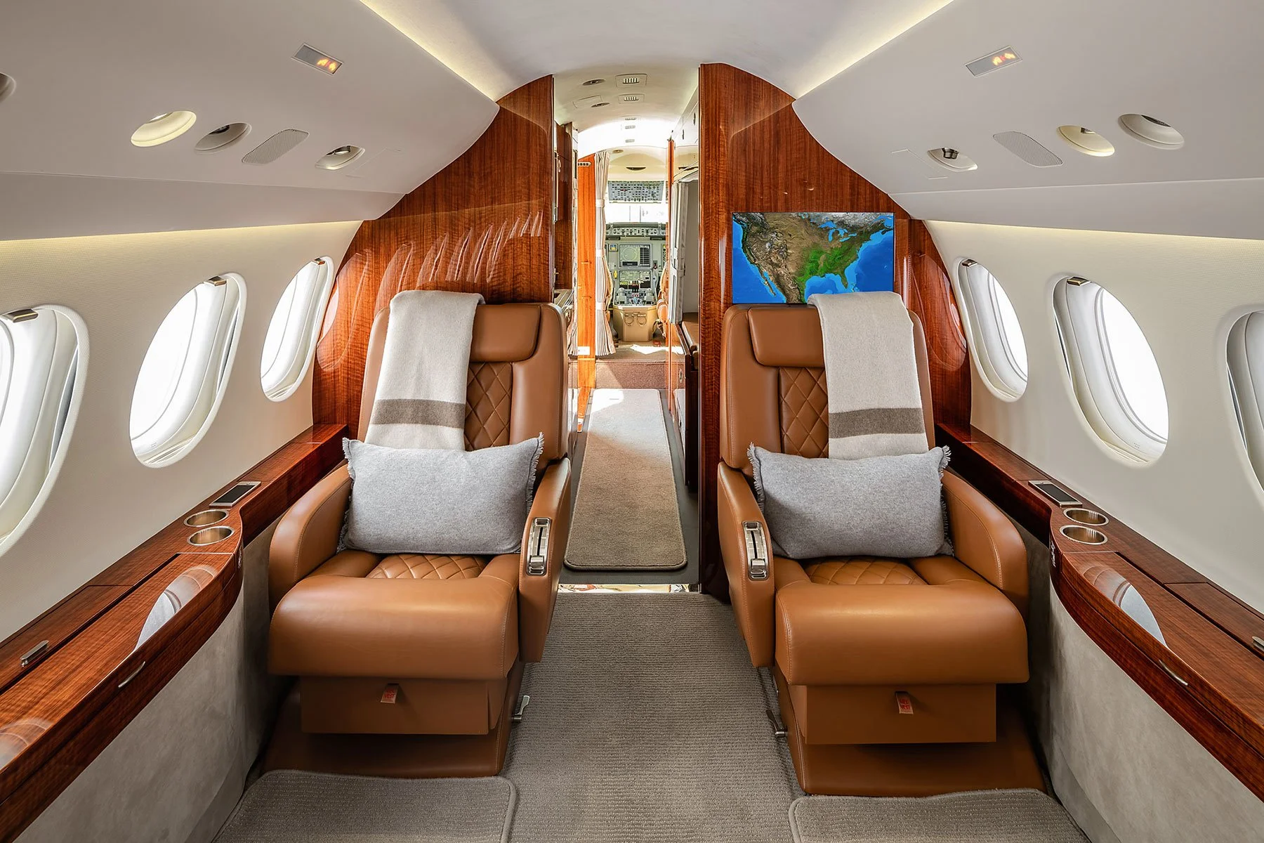 Inside the luxurious cabin of a private jet, showing two brown leather seats with gray cushions, wood paneling, and a flat-screen TV displaying a map. The cabin has small oval windows with blinds and an aisle running through the middle, leading to the cockpit at the front.