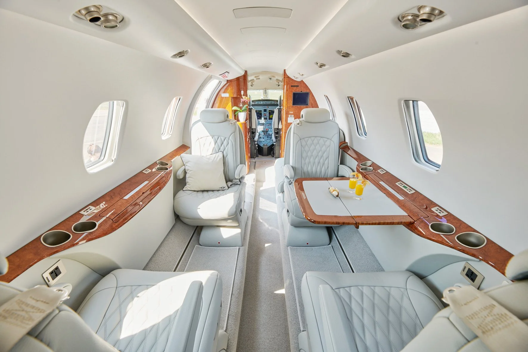 Interior of a private jet with luxurious white leather seats, wooden accents, and windows on both sides, with a view of the cockpit at the front.