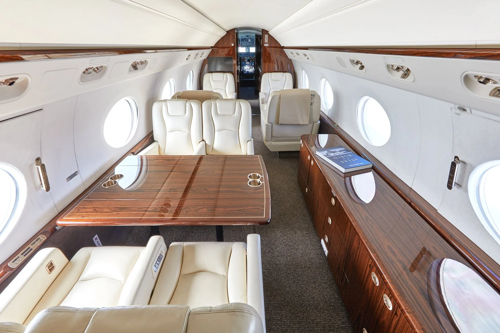 Interior of a private jet with cream leather seats, wood paneling, and a large wooden table with cup holders, with windows along the sides and a cockpit visible at the front.