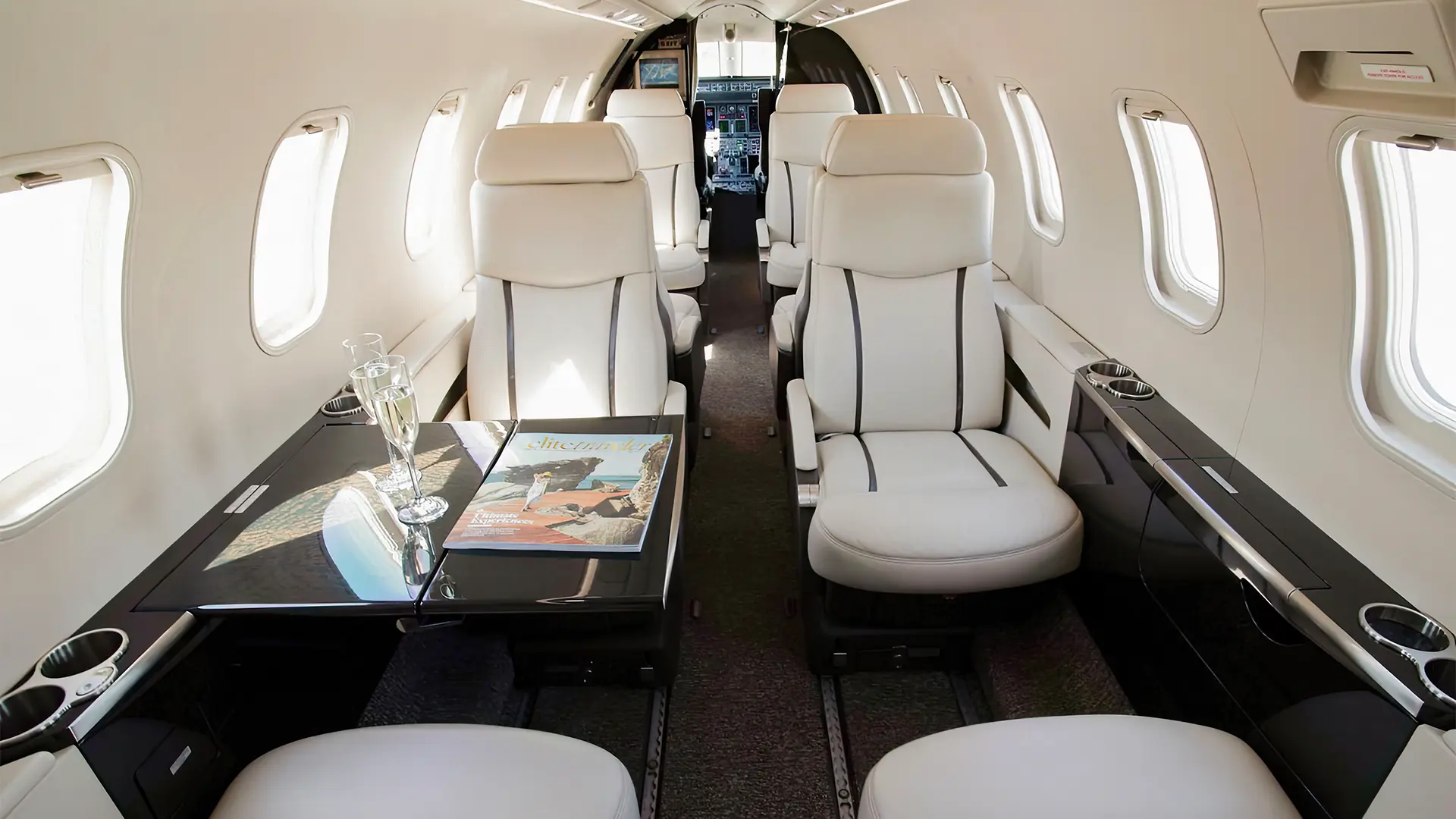 Interior of a private jet with white leather seats, black glossy tables, and windows on both sides. There are two glasses of champagne and a magazine on the table in the foreground.