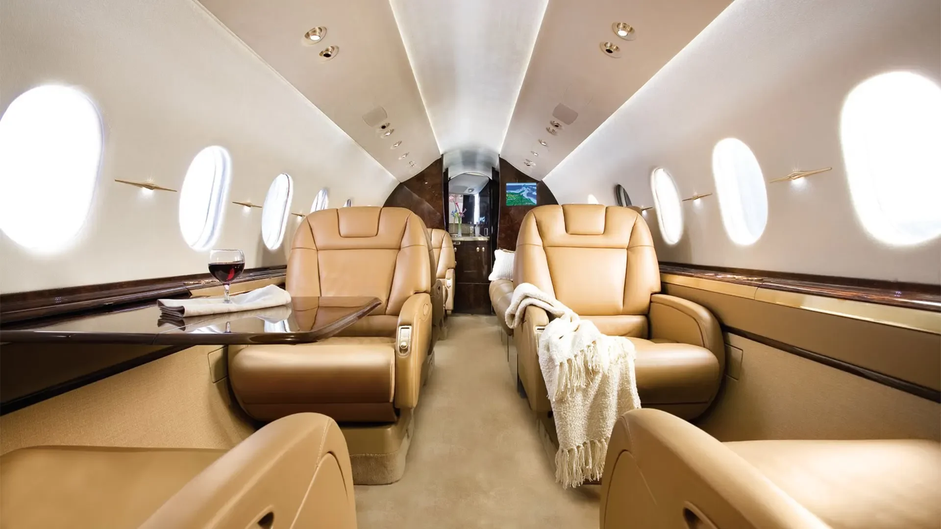 Interior of a private jet with beige leather seats, a table with a glass of red wine, and windows along the walls.