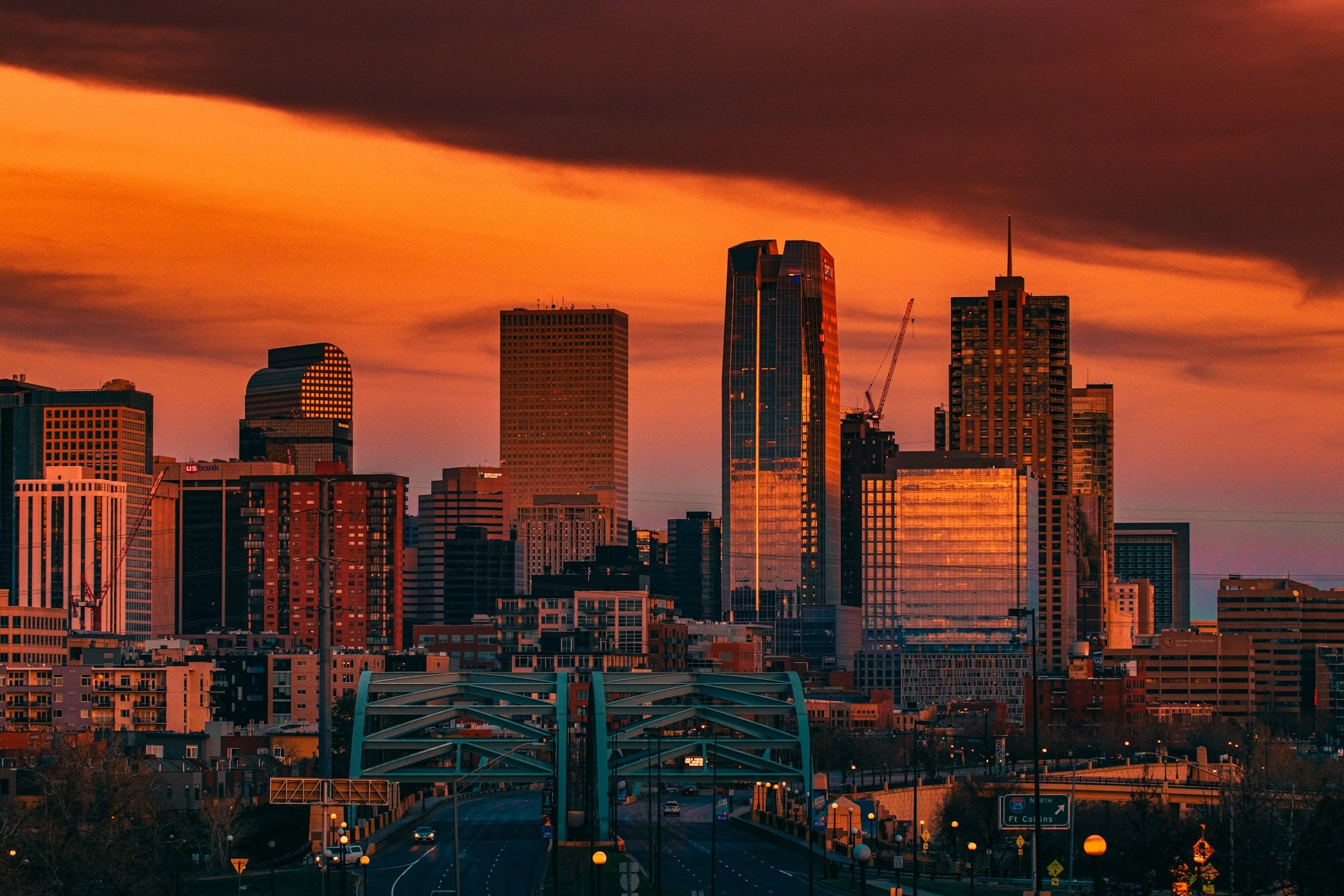 Sunset view of a city skyline with tall skyscrapers and a bridge in the foreground, under a dramatic, colorful sky.