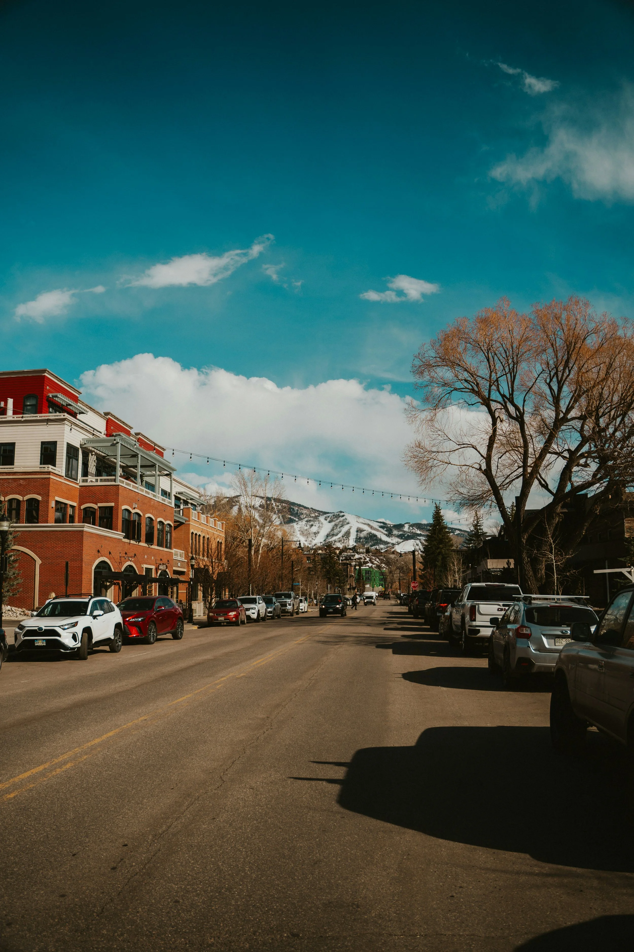 A street scene in town with parked cars, trees, buildings, mountains in the background, and a partly cloudy sky.
