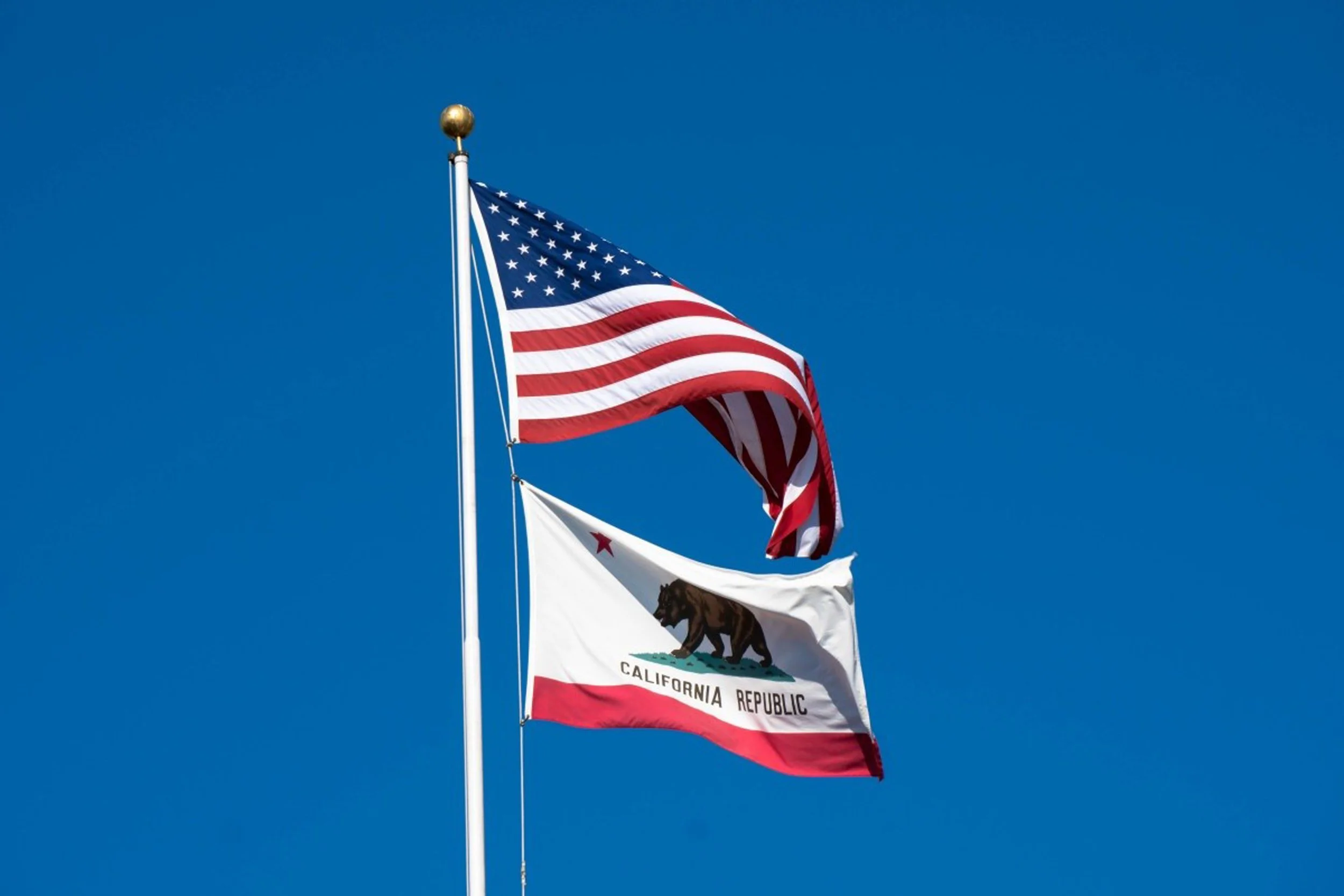 Two flags on poles against a clear blue sky: the American flag with red, white, and blue stripes and stars, and the California state flag with a bear.