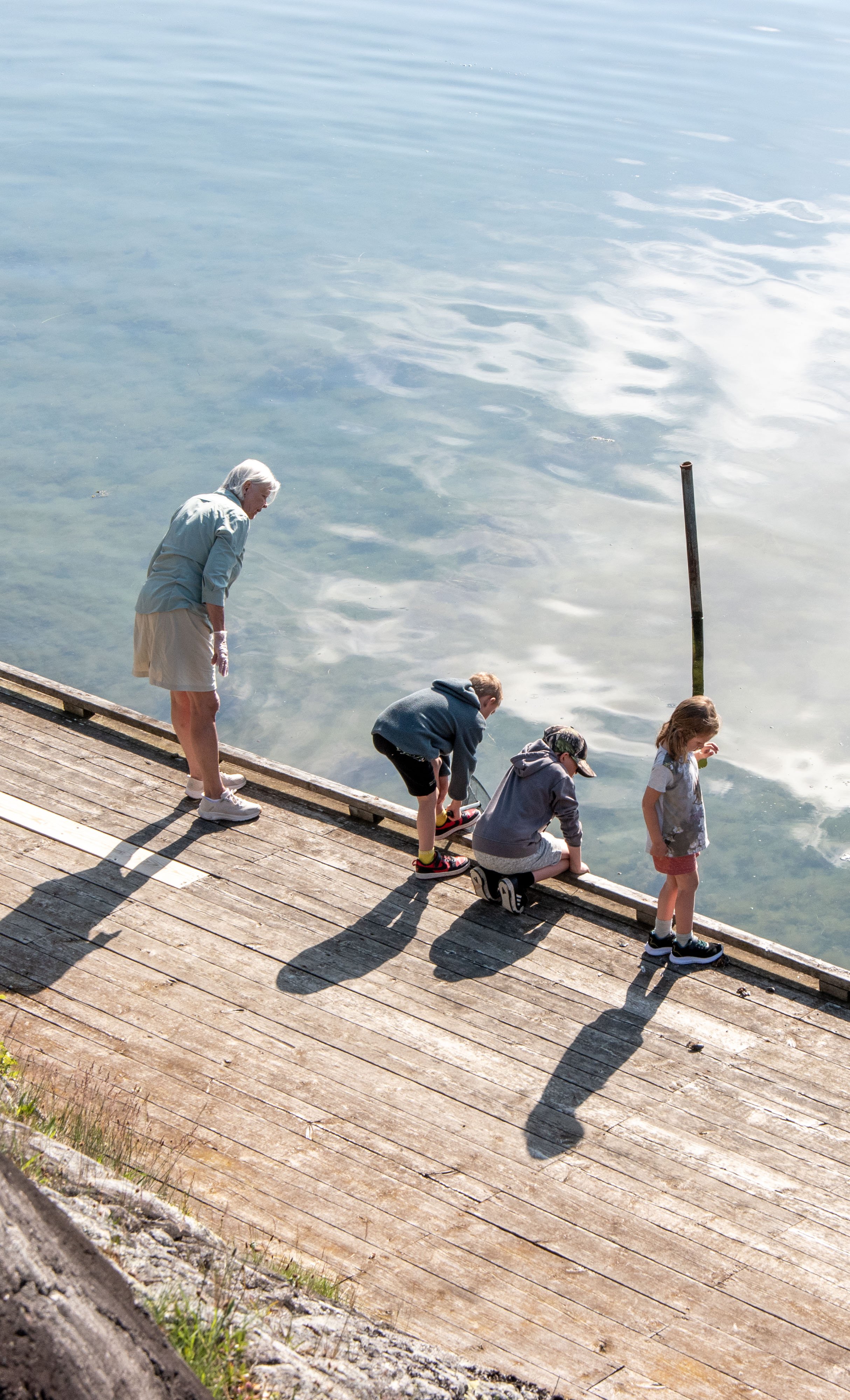En äldre kvinna och tre barn står på en vedbrygga vid vattnet. Barnen tittar ner i vattnet, och kvinnan står bredvid dem.