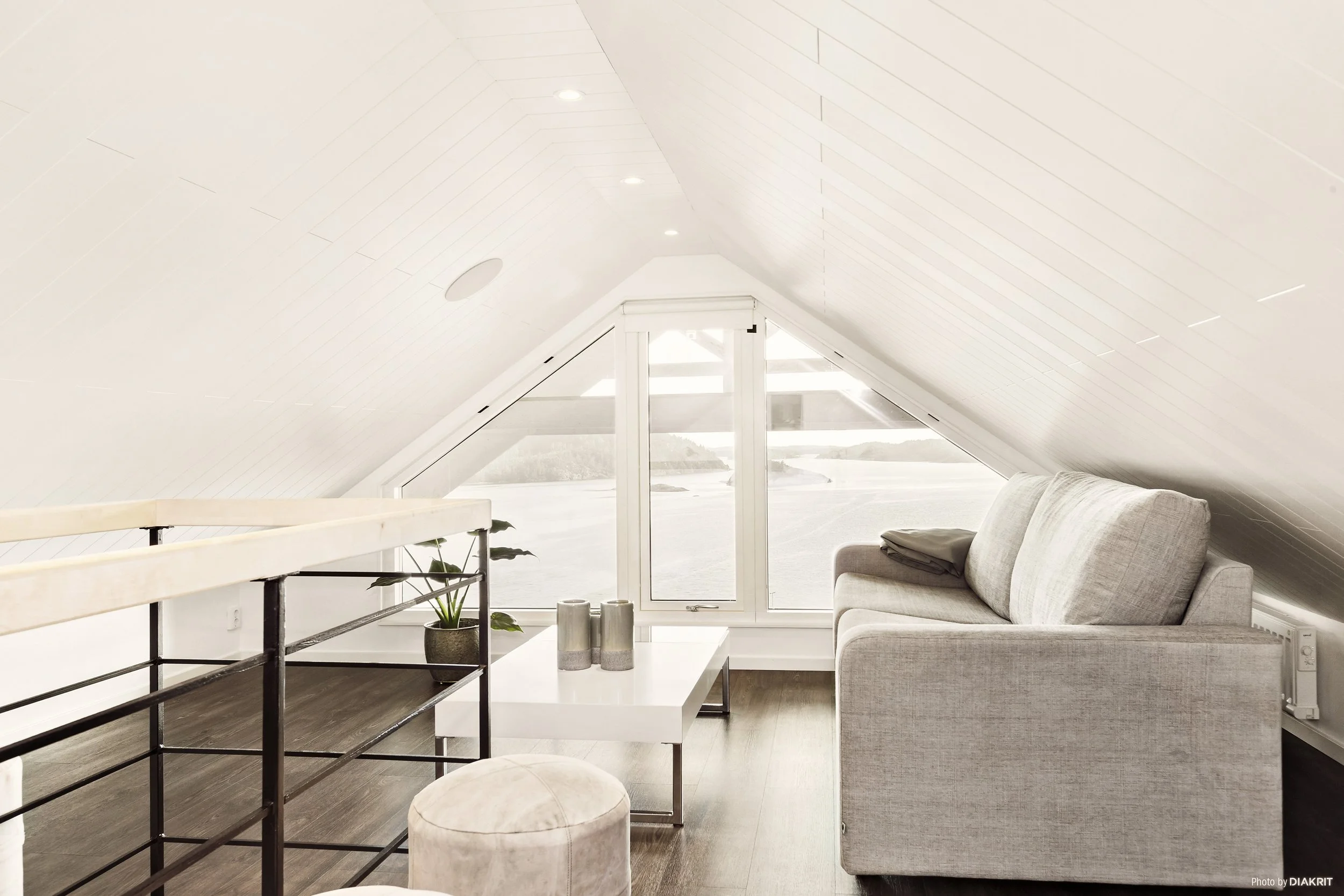 Rymligt vardagsrum med en soffa, ett soffbord, en växt och stora fönster med utsikt över vattnet. Väggen och taket är vit och sluttande.