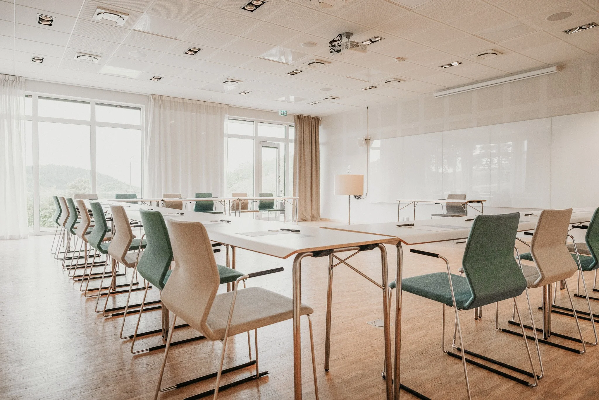 Une salle de classe vide avec une table de conférence et des chaises aux couleurs claires, de grandes fenêtres avec des rideaux blancs et un tableau blanc au mur.