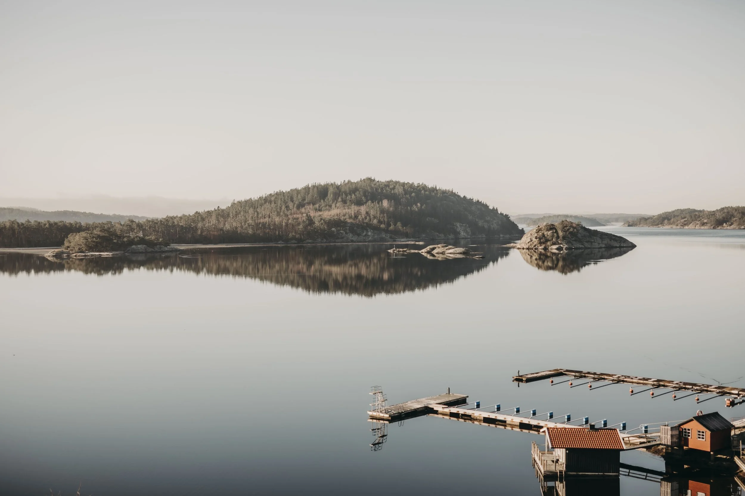 En lugn och stilla sjö med reflektion av en ö med skog, samt en liten brygga och en liten byggnad i förgrunden.