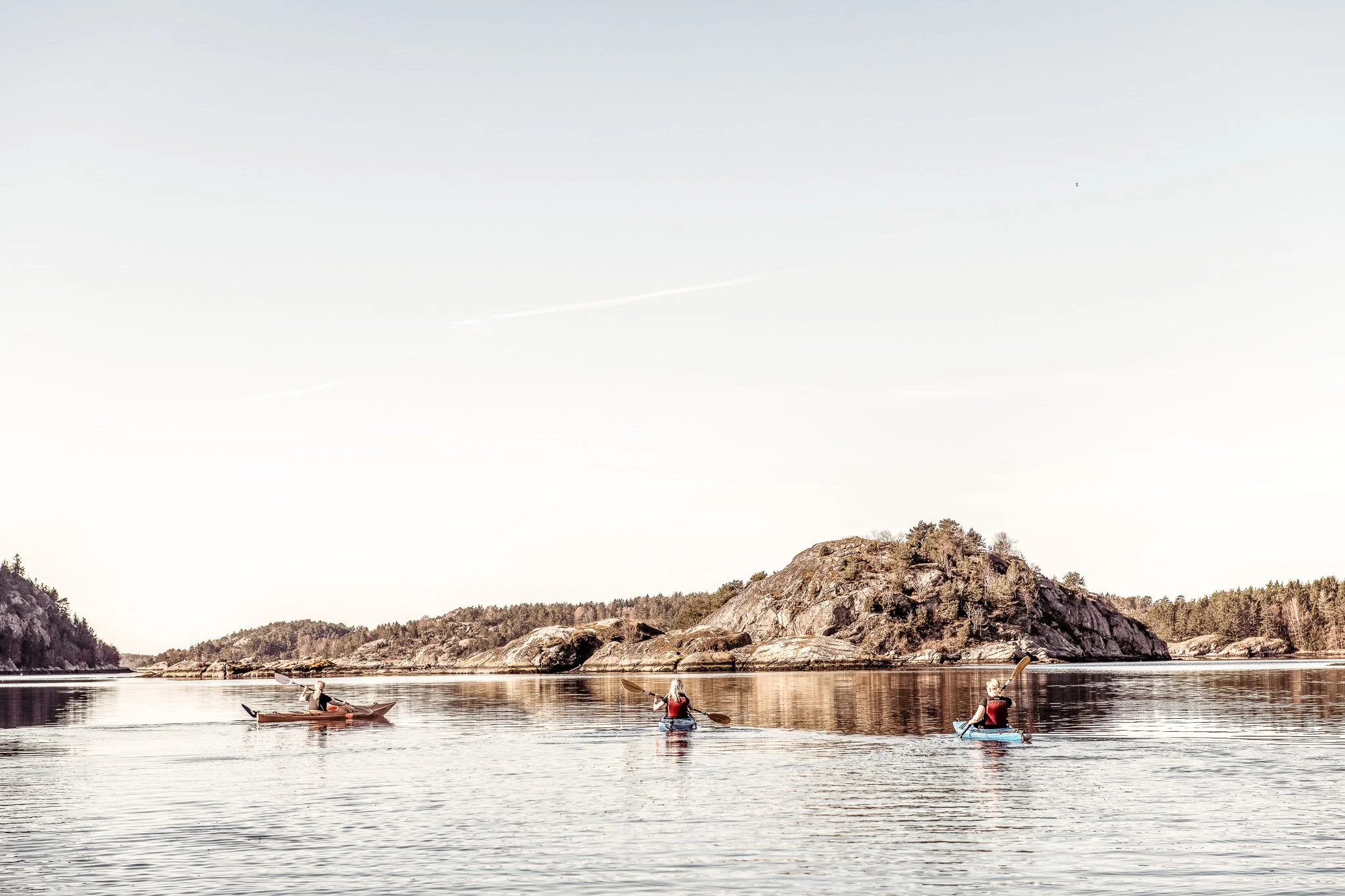 Tre personer paddlar kanot på en stilla sjö med en ö i bakgrunden, omgiven av skog och klippor, under en klar himmel.