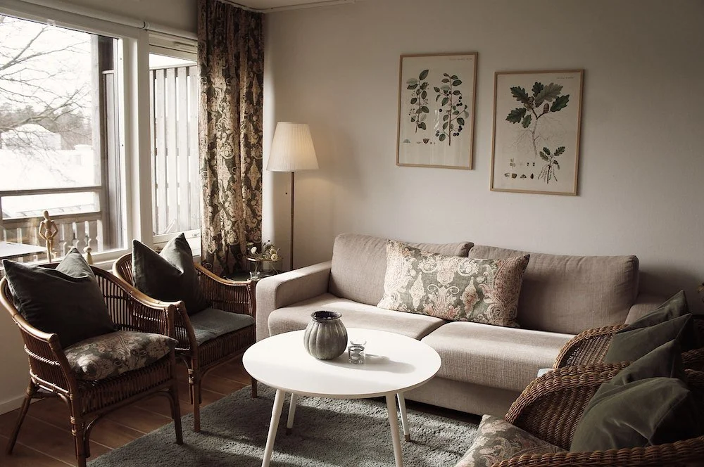 Vardagsrum med stor soffa, fåtöljer med kuddar, ett soffbord med vaser, väggdekorationer med bladmotiv, en golvlampa, fönster med gardiner och en skymtande utsikt.
