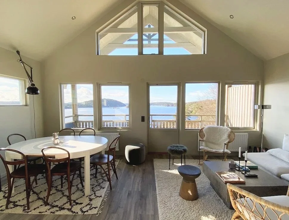 Vardagsrum med stora fönster och utsikt över vattnet, matbord med sex stolar, soffor, fåtöljer och bord, ljusa väggar och trägolv.