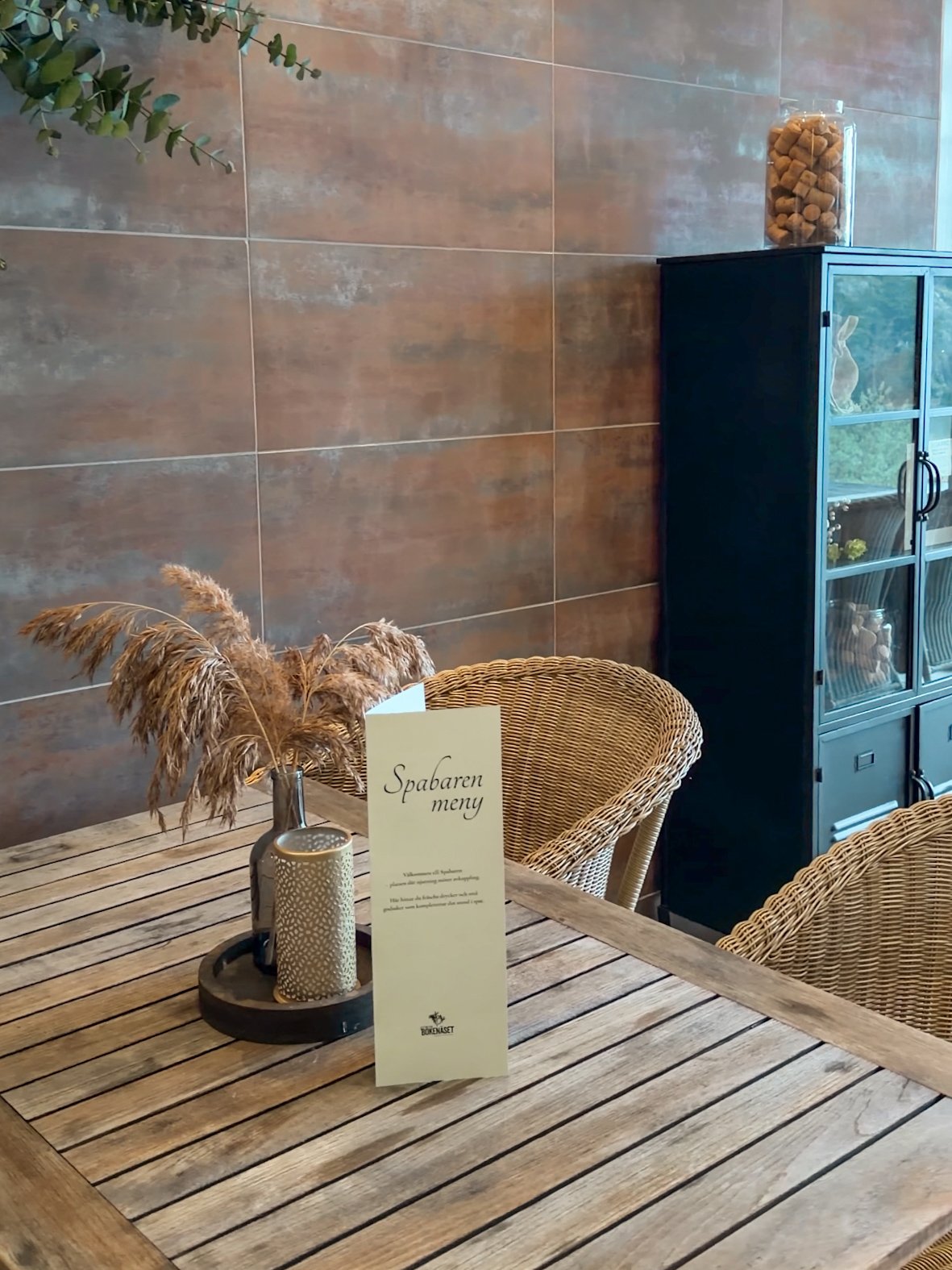 Rustikt träbord med vaser och ett menykort, omgiven av rottingstolar, i ett café eller restaurang med tegelväggar och ett skåp i mörk färg.