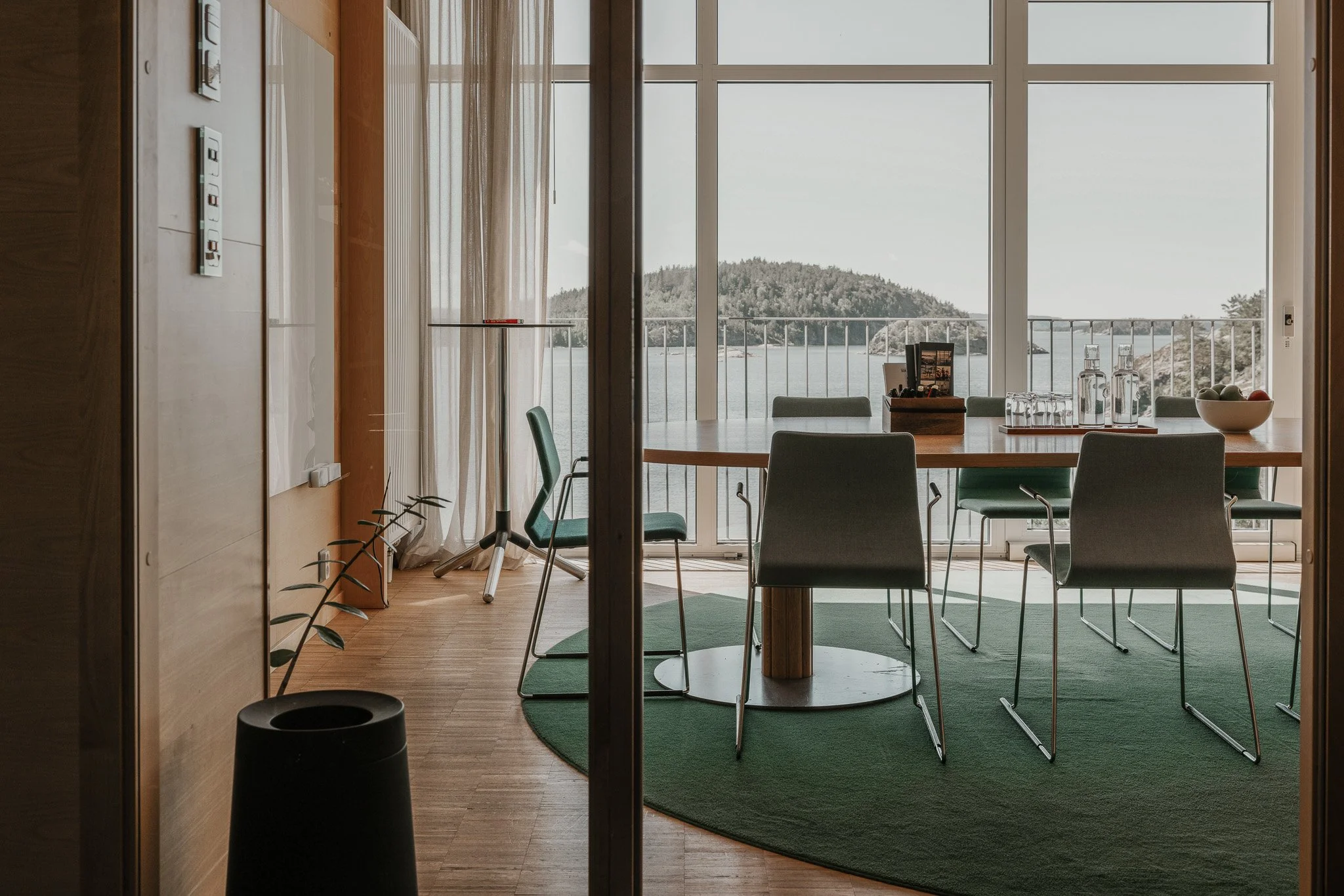 Une salle de conférence avec de grandes fenêtres donnant sur la mer et les îles au large. Une table ronde en bois avec six chaises vertes, quelques bouteilles d'eau et un bol de fruits sur la table. La pièce est dotée d'un parquet et d'un espace vert sous la table.