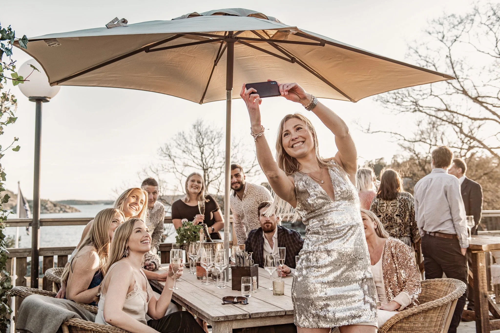 En grupp människor firar utomhus under en stor parasoll, där en kvinna i en silverklänning tar en selfie med andra som sitter och står runt ett bord med drycker.